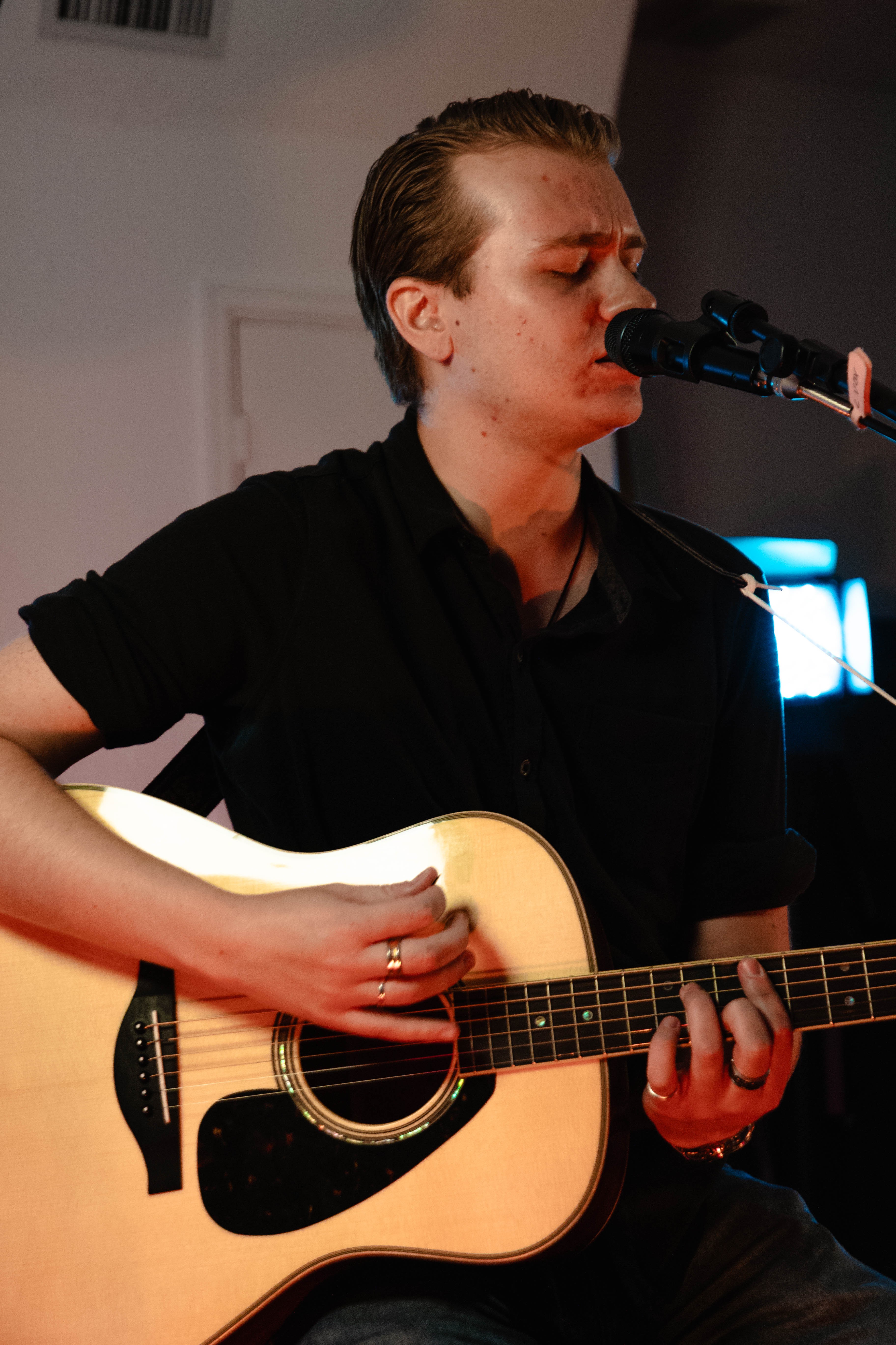 Person with short hair playing an acoustic guitar and singing into a microphone.