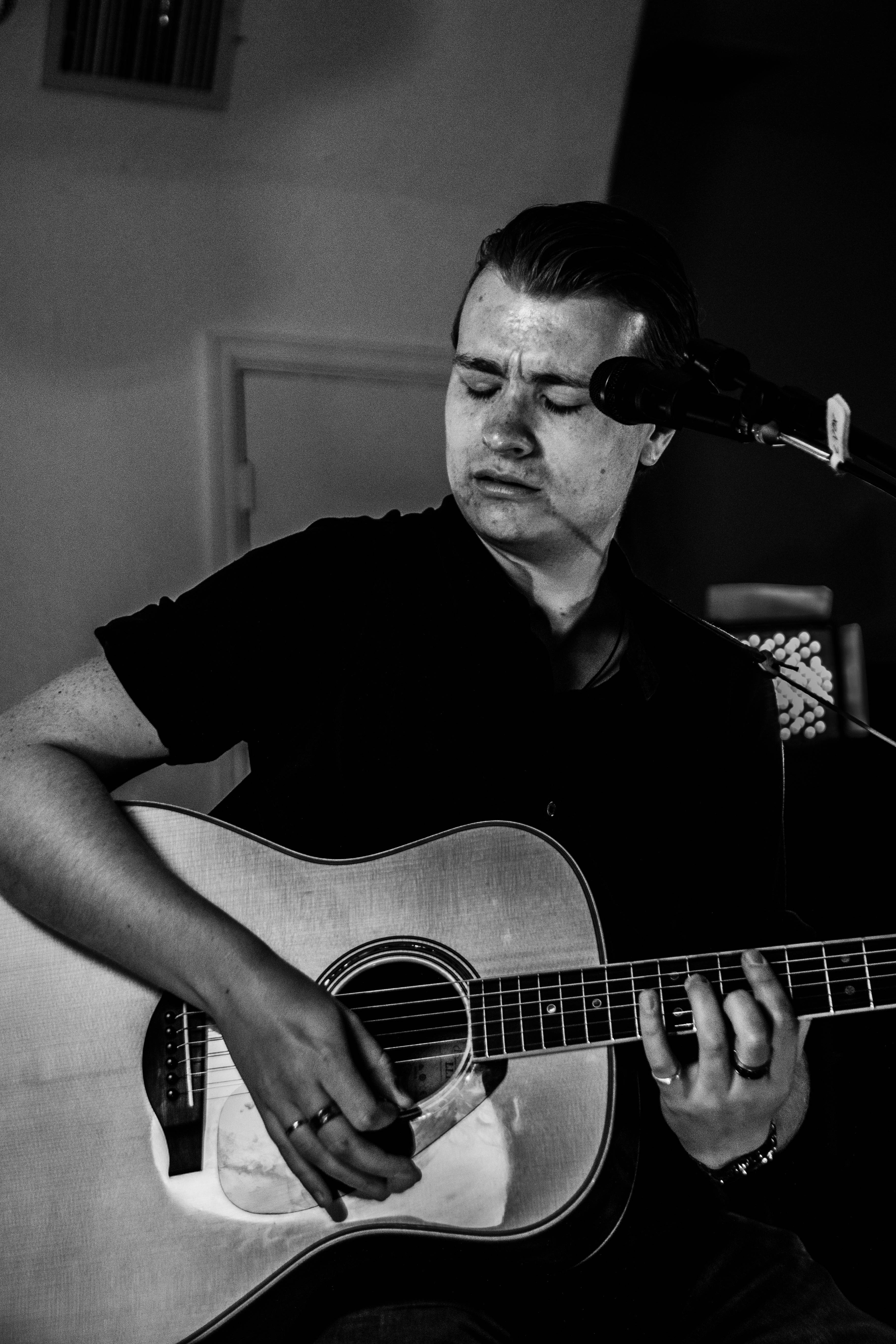 Person playing an acoustic guitar with eyes closed, microphone nearby, in a dimly lit room.