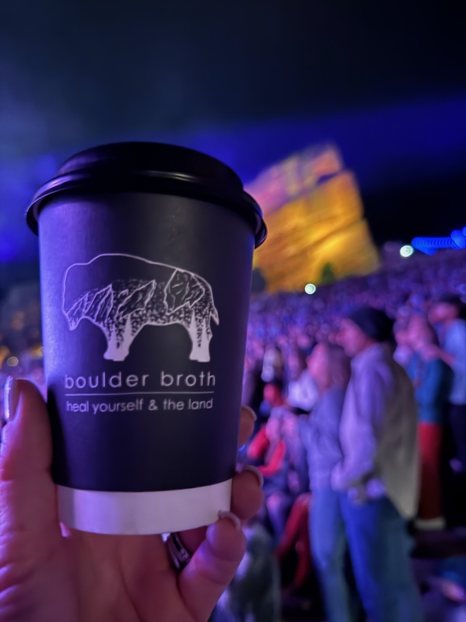 Hand holding a black coffee cup with a bison logo and text, crowd and stage in background at night.