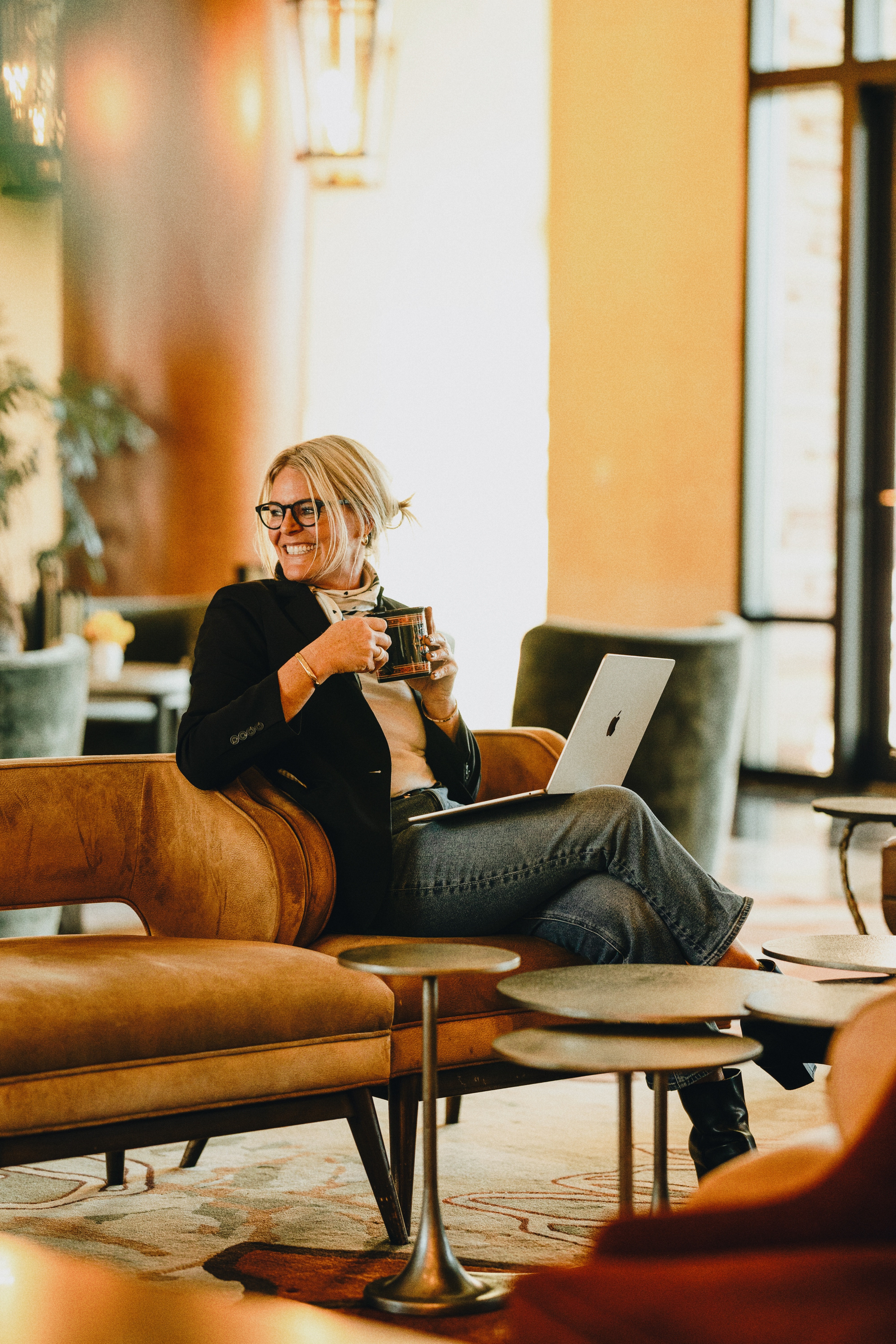 Woman with glasses sitting on a sofa, holding a mug, with a laptop nearby, in a bright cafe setting.