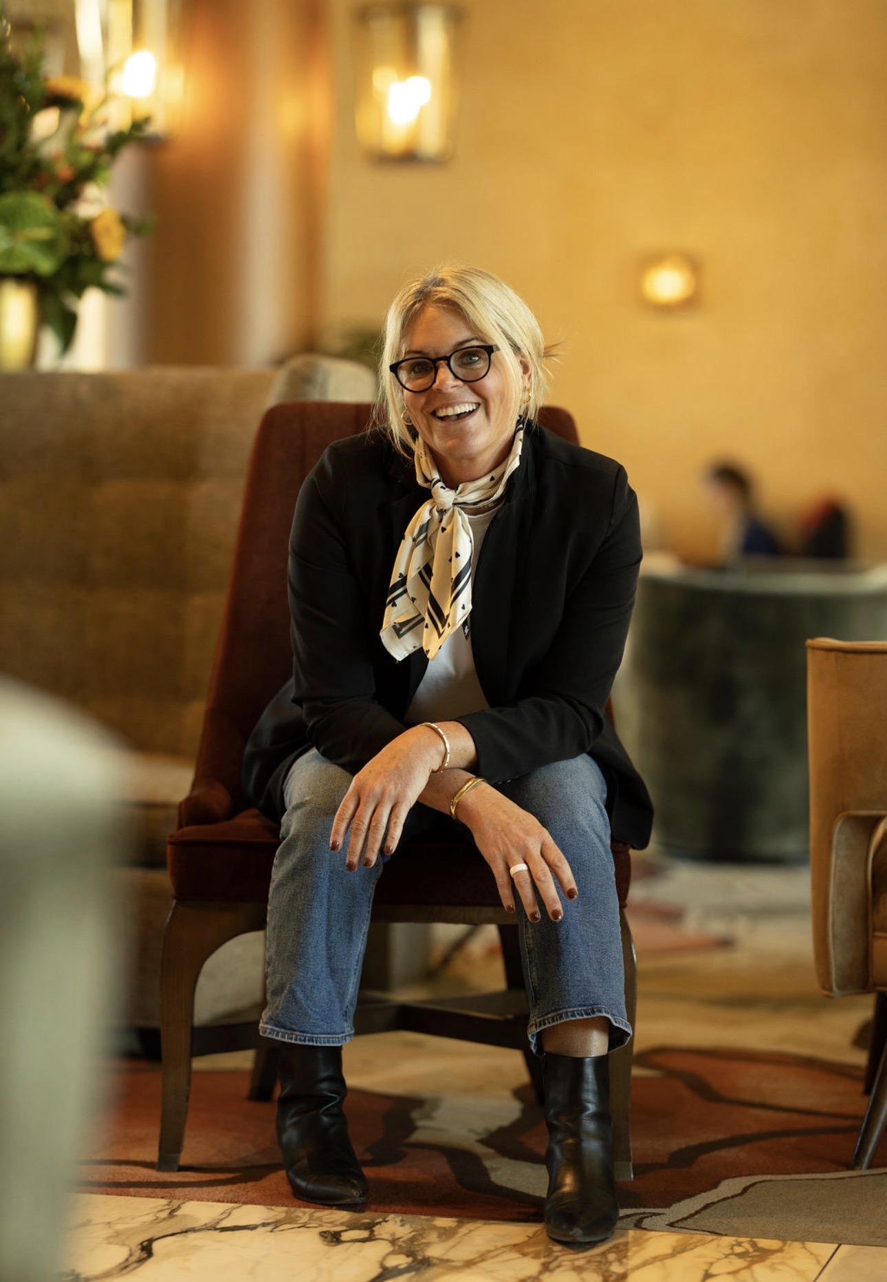 Woman with glasses and blonde hair sitting on a chair, smiling, in a warmly lit indoor setting.