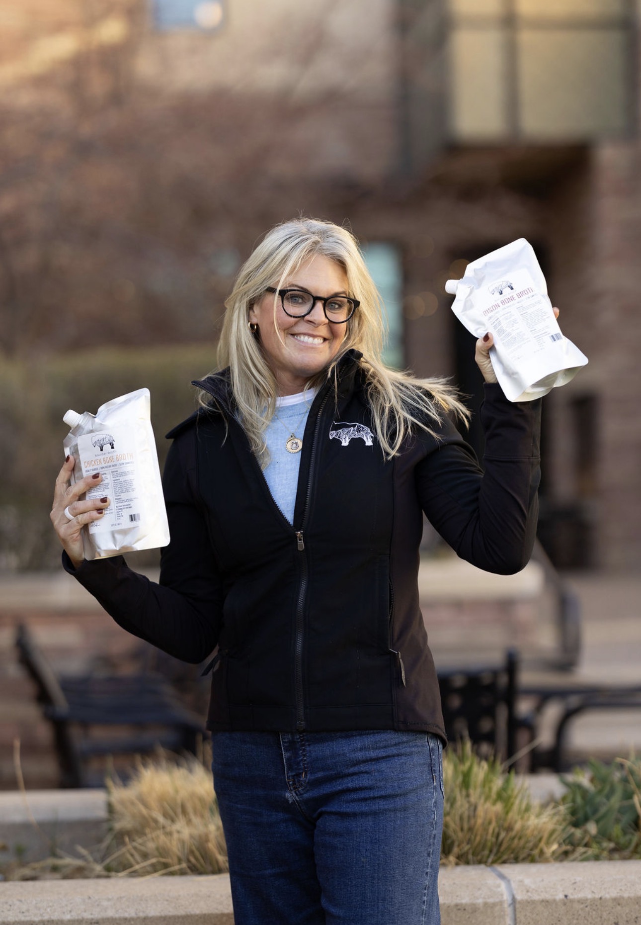 Woman with glasses smiling outdoors, holding paper bags in both hands, wearing a black jacket and blue jeans.