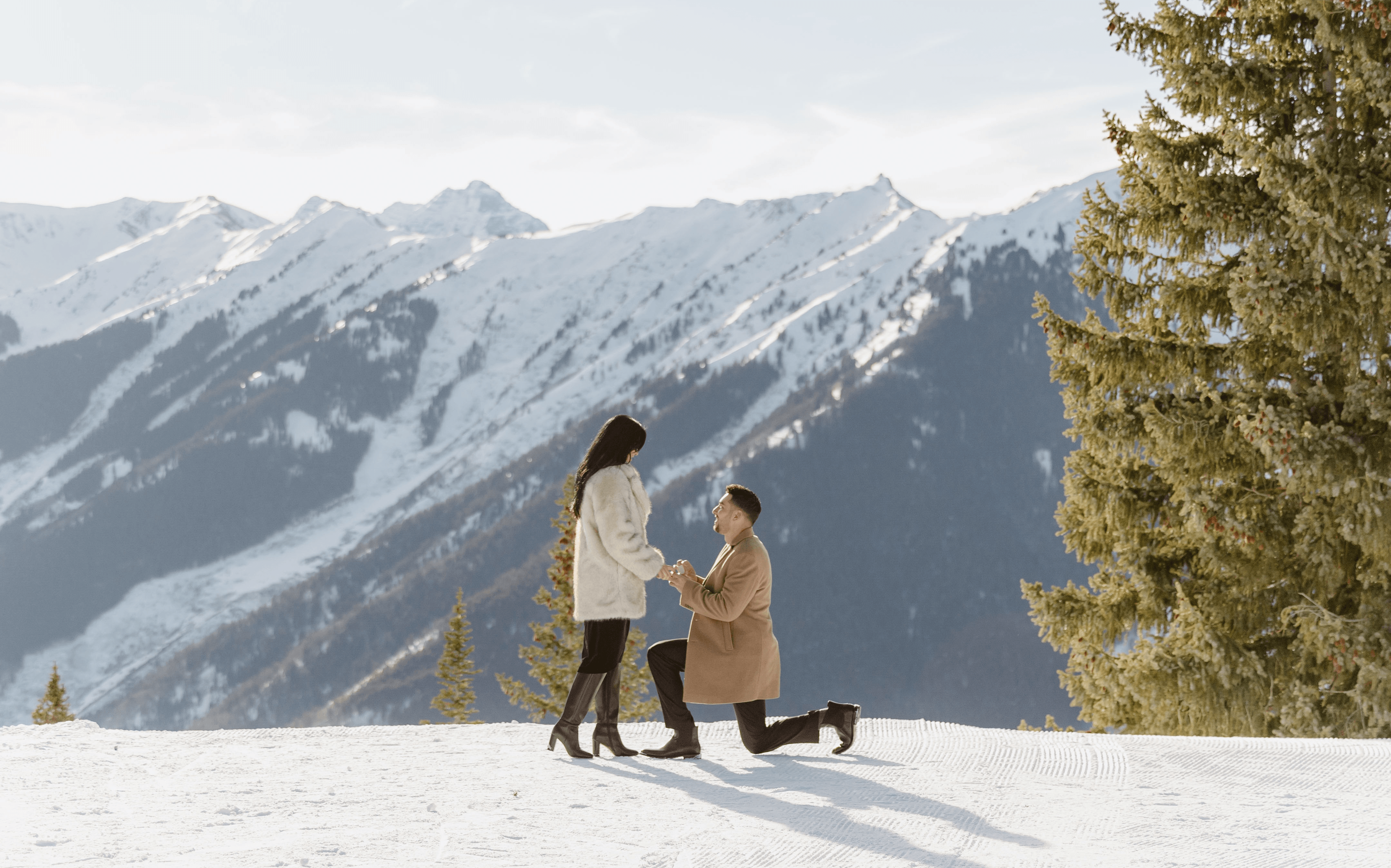 Two people, one kneeling, exchange rings outdoors with snow-covered mountains in background.