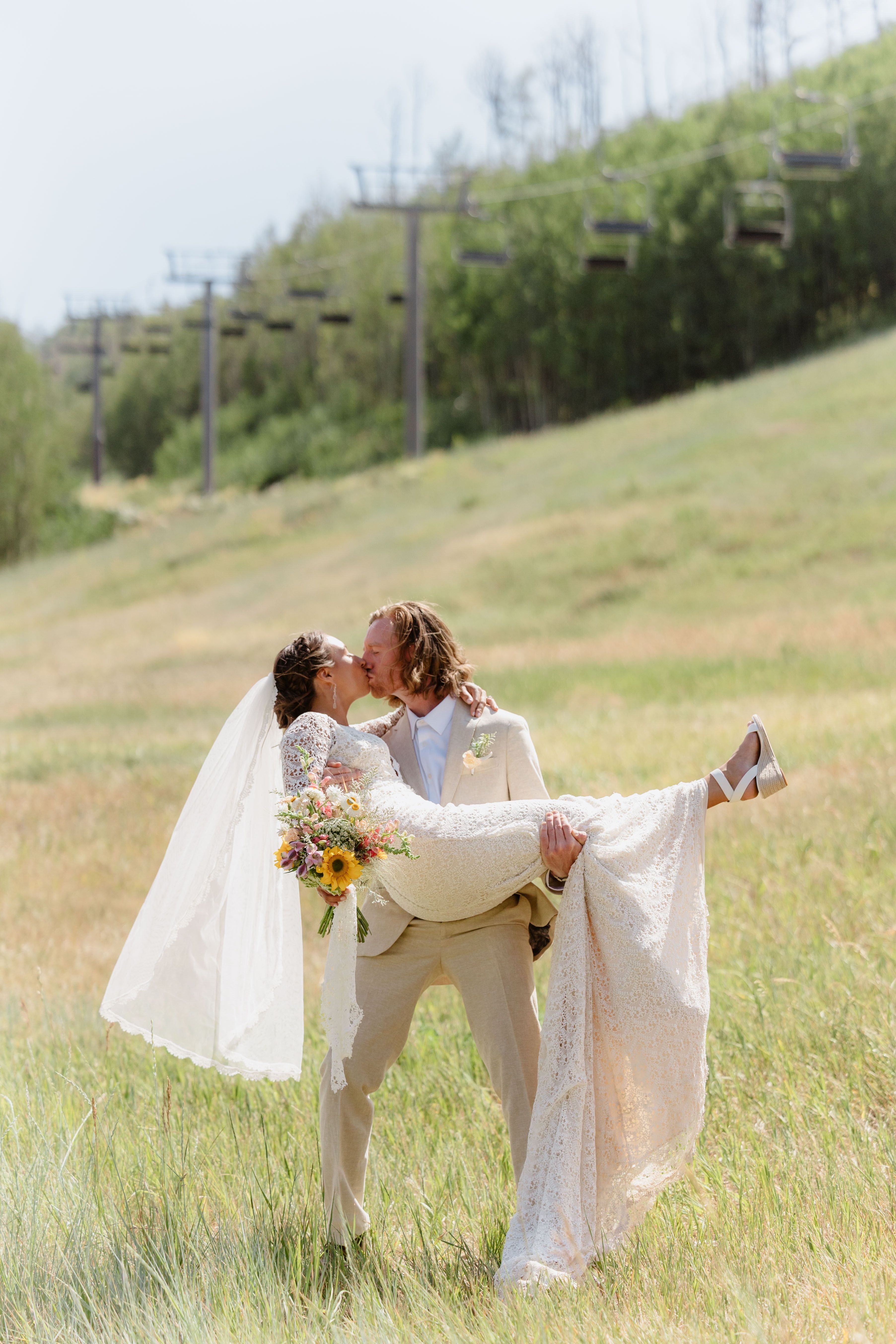 A couple in wedding attire sharing a kiss outdoors on a grassy field with trees and power lines in the background.