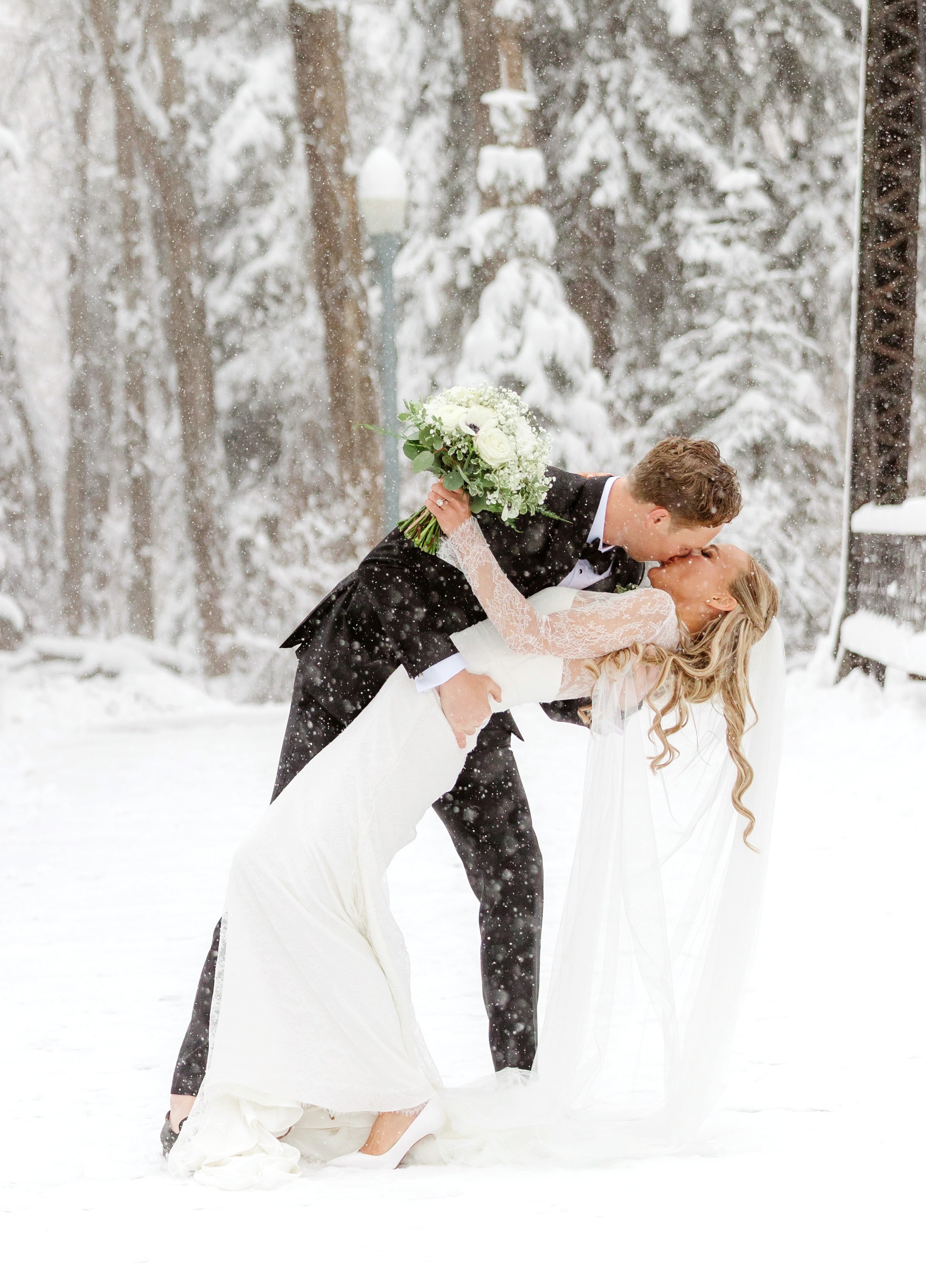 A couple in wedding attire sharing a kiss outdoors in a snowy forest, with trees and snow in the background.