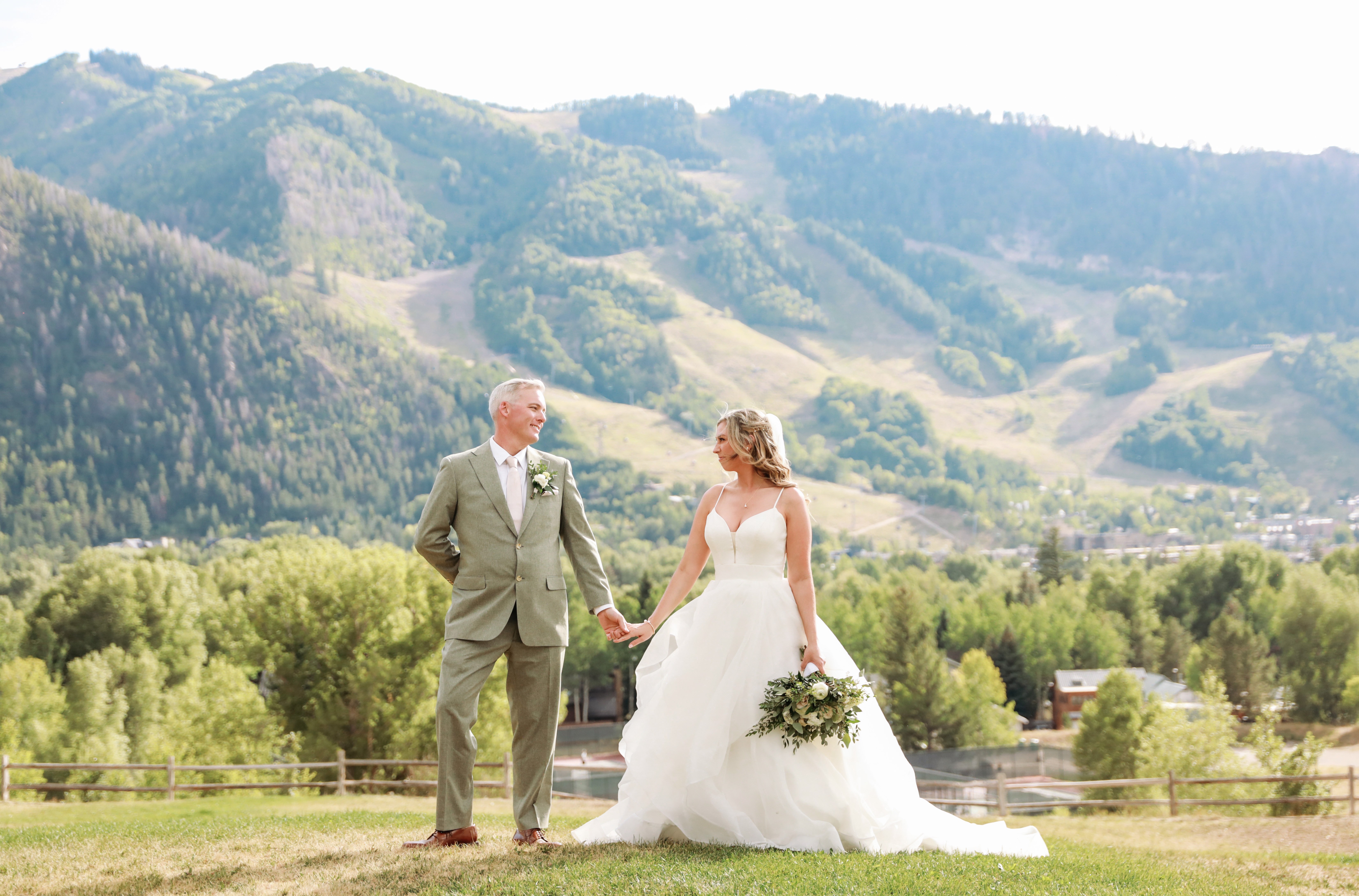 A bride and groom holding hands outdoors with mountains and greenery in the background.