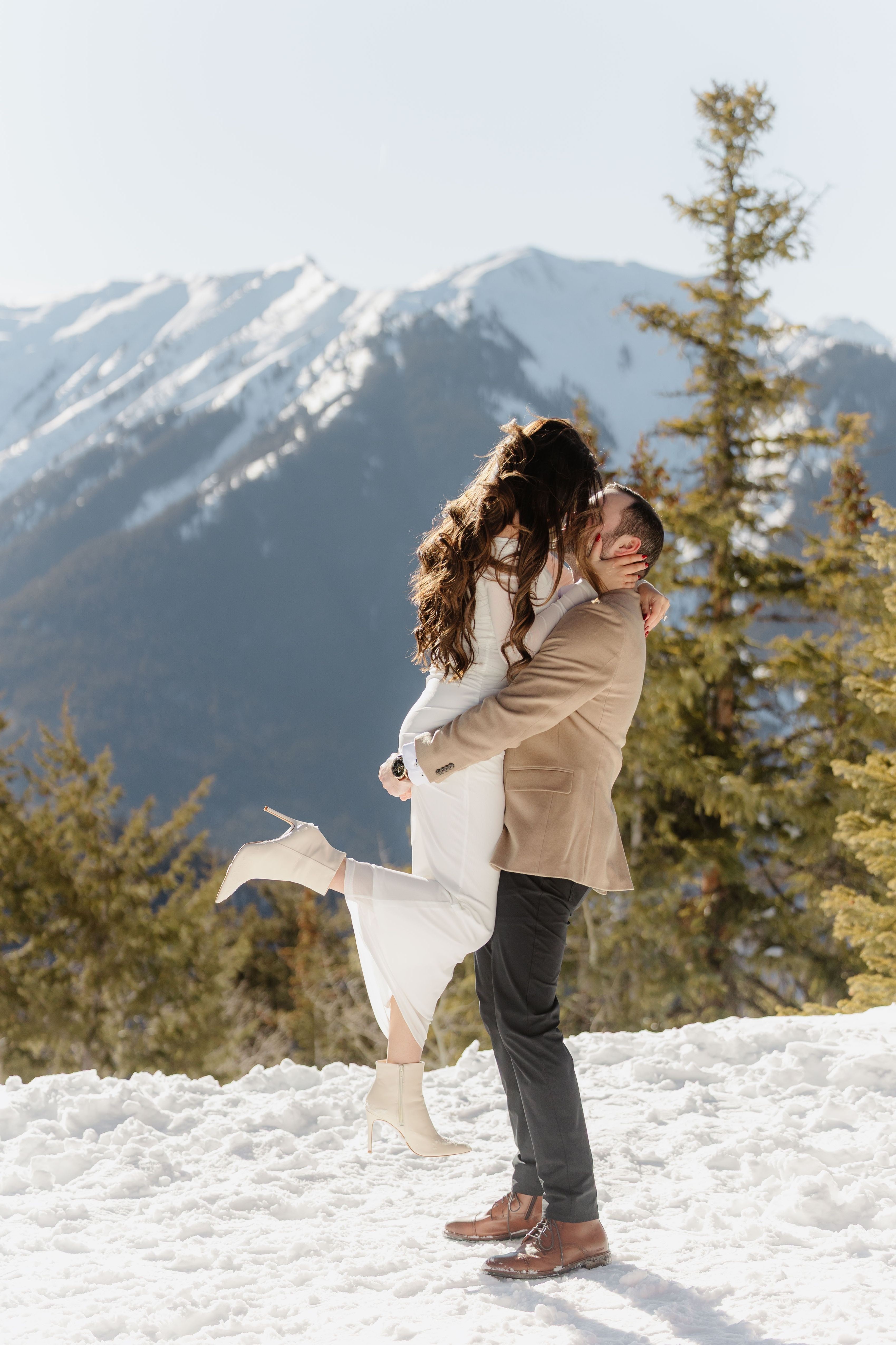 A couple walking in a snowy landscape with mountains and trees in the background, embracing each other.