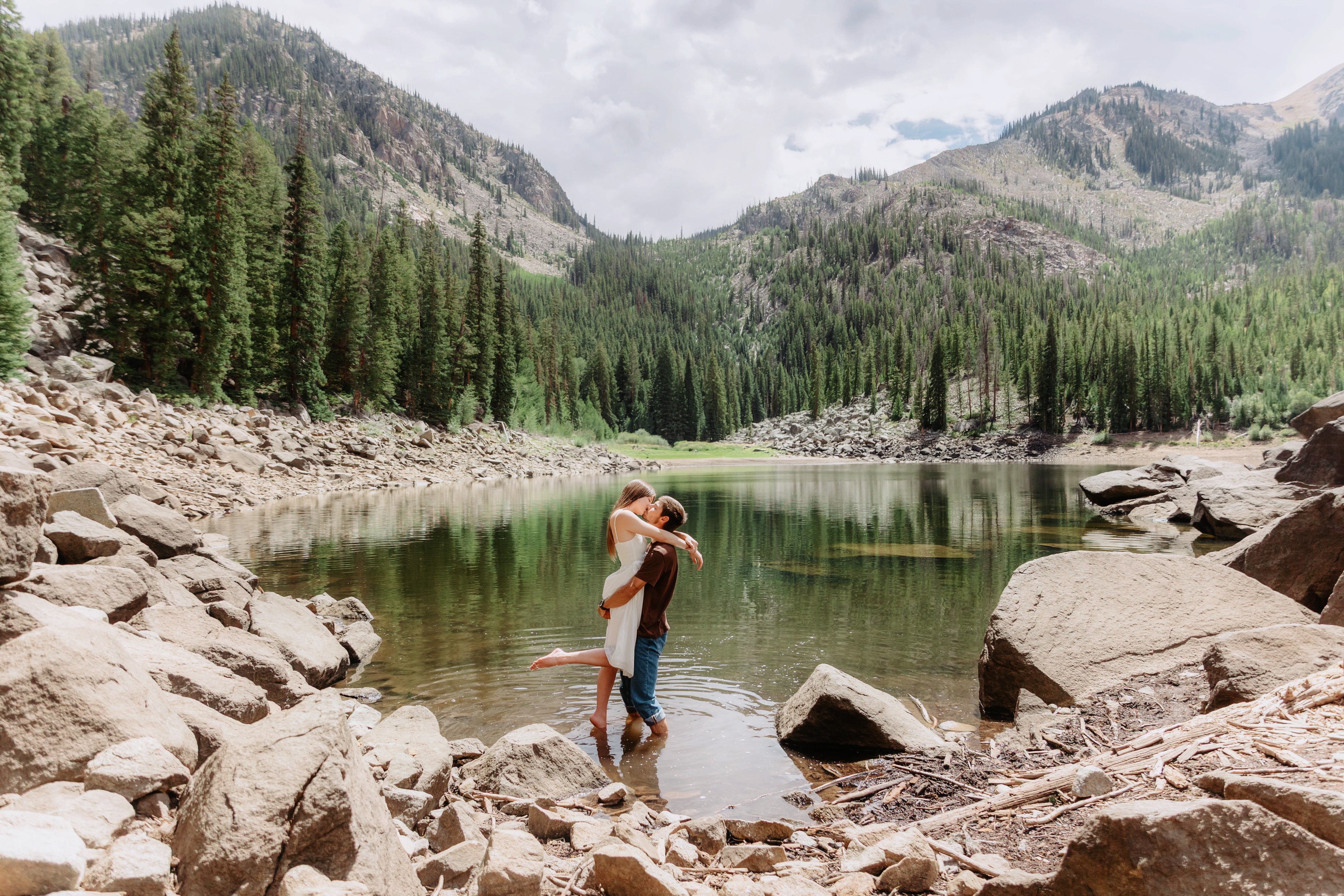 Two people stand on rocks by a lake surrounded by mountains and trees, with cloudy sky above.