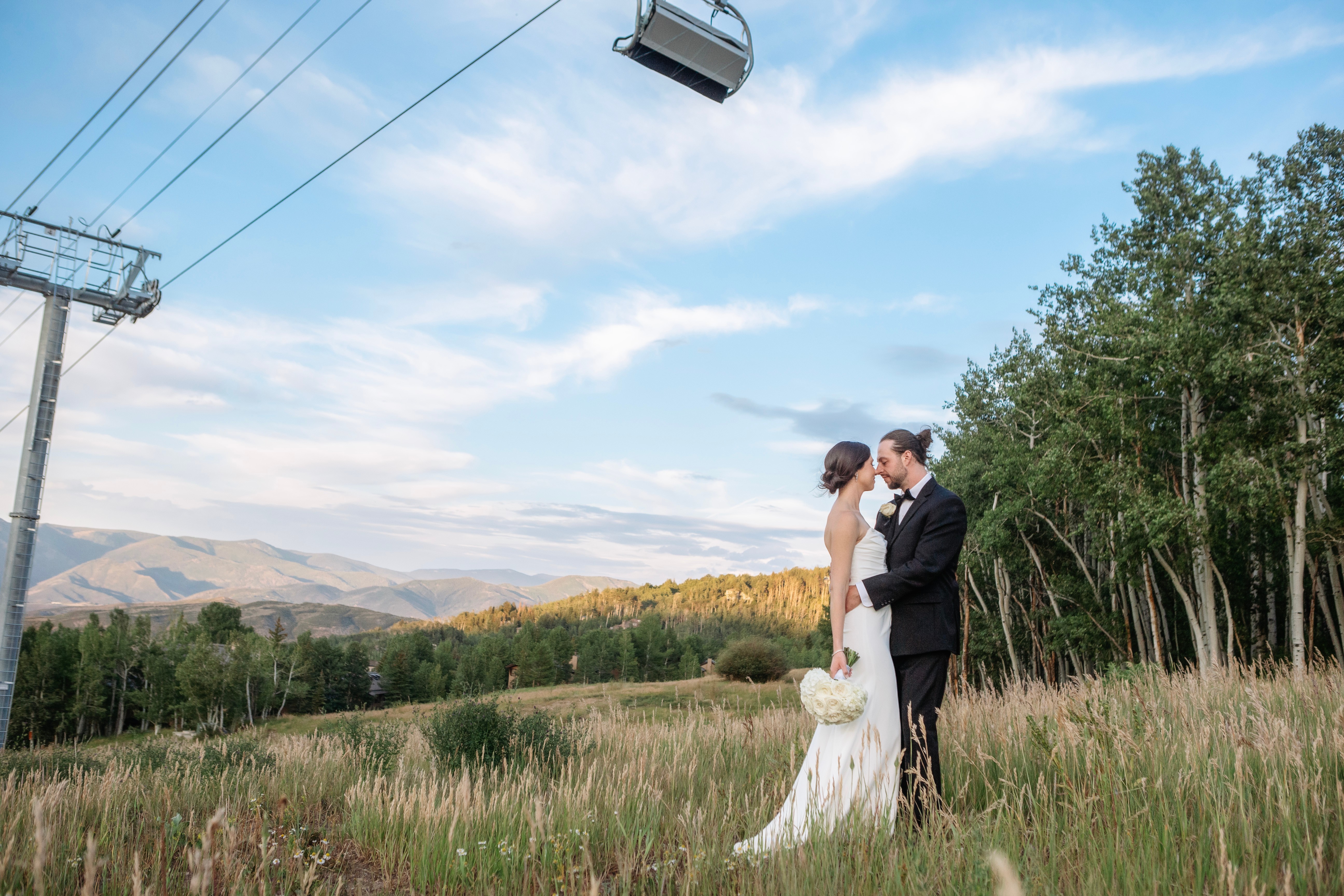 A bride and groom stand close together in a grassy field, holding hands, with trees and mountains in the background.