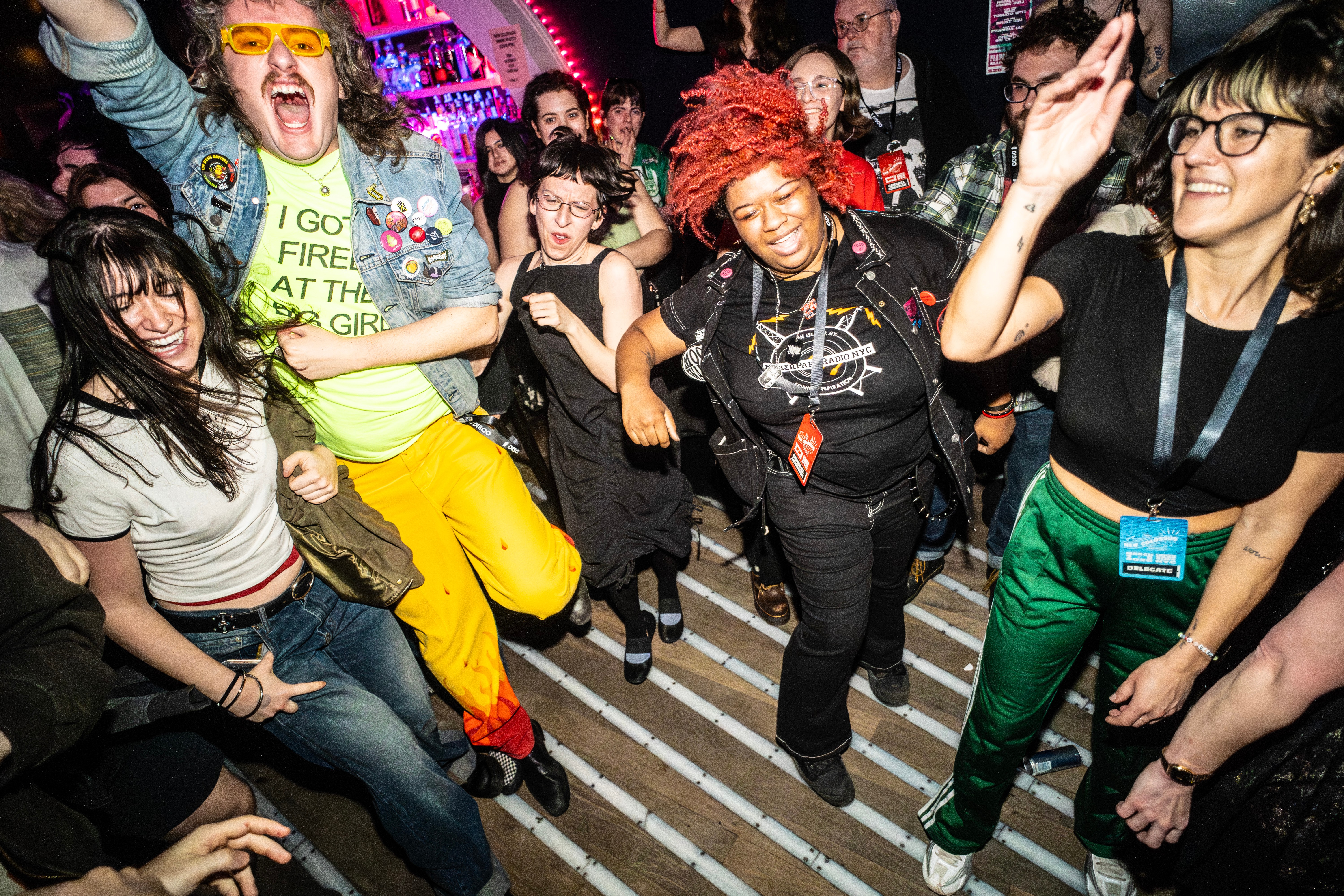 Group of people dancing and enjoying a party with colorful lighting, some smiling and raising hands, in a lively indoor setting.