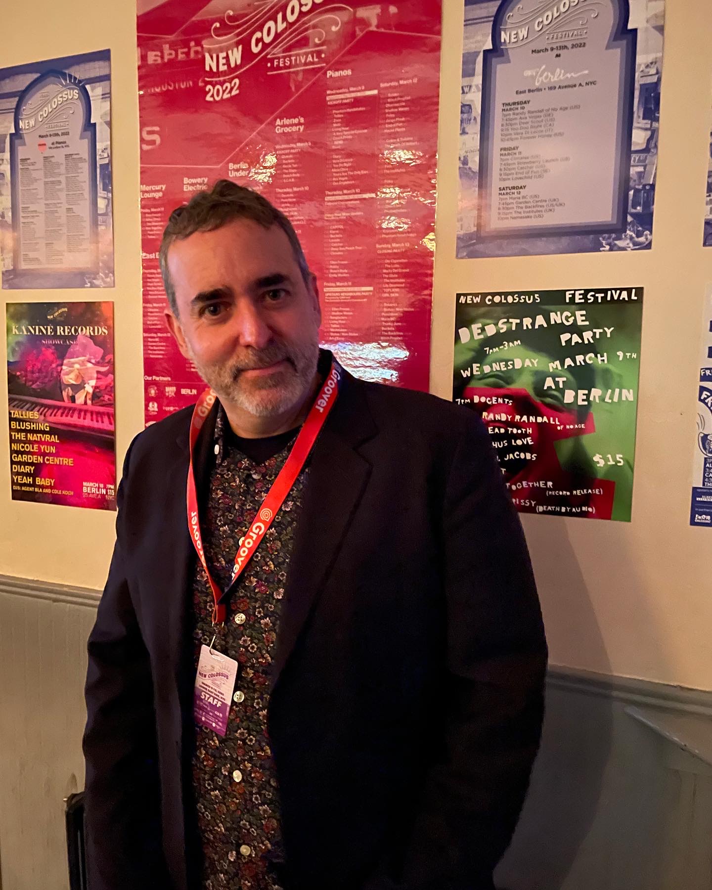 Man with short dark hair and beard wearing a black jacket and patterned shirt, standing in front of colorful posters on a wall.