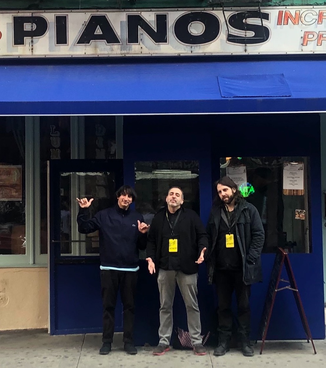 Three people standing outside a store with a sign that reads 'PIANOS'. They are wearing dark jackets and have yellow badges.