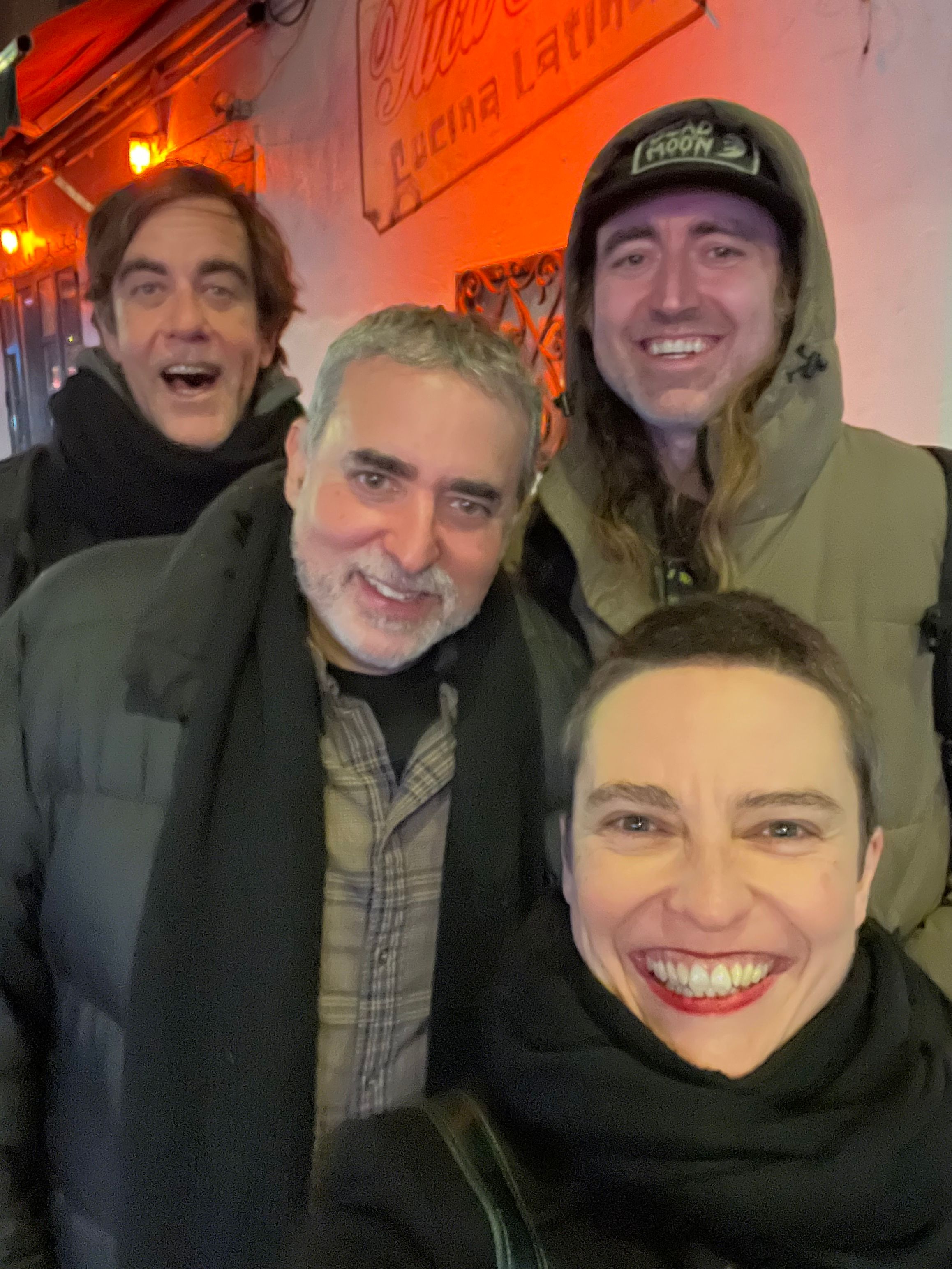 Four people smiling and taking a selfie indoors with warm lighting.