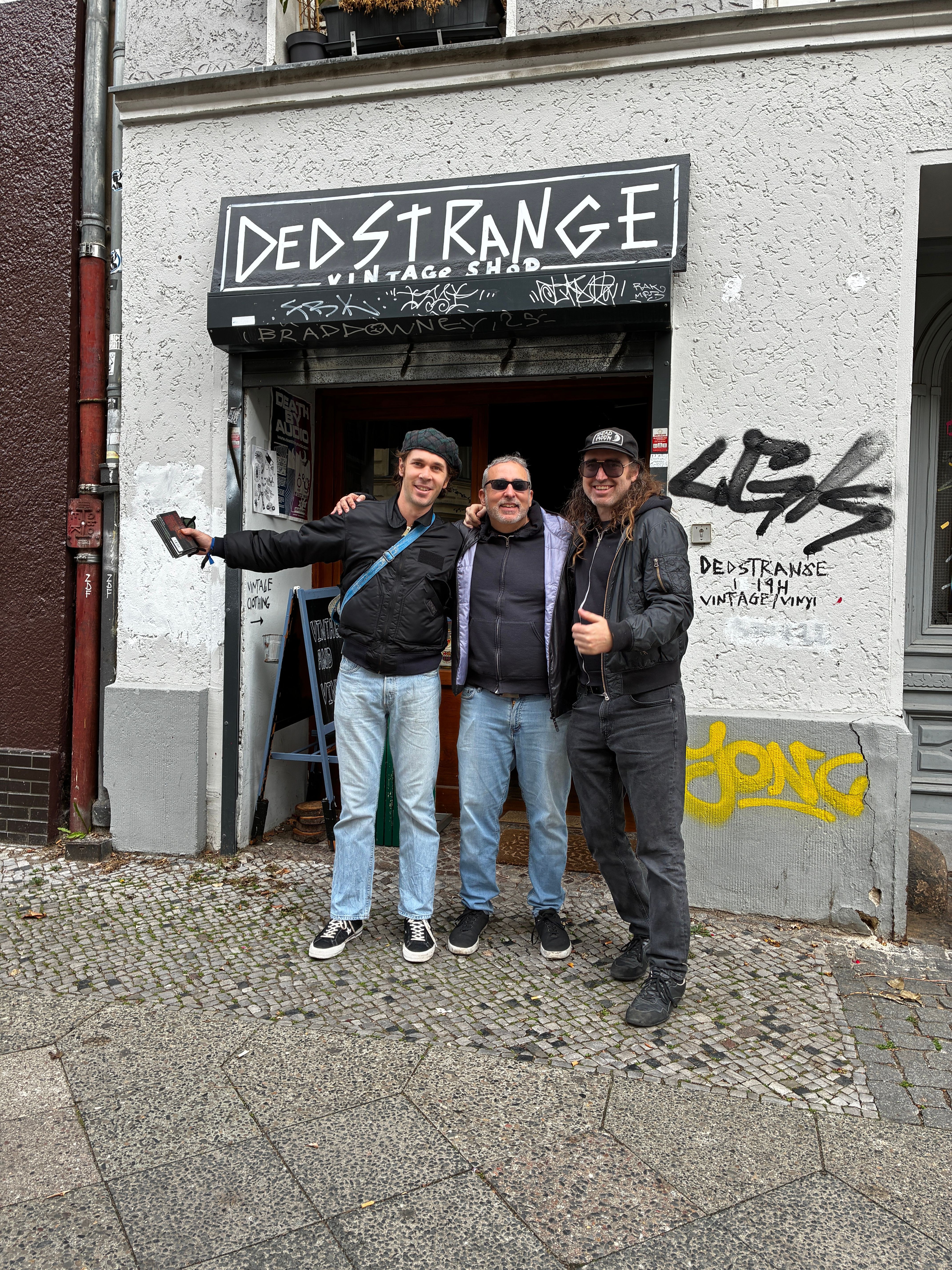 Three people stand outside a building with a sign reading 'DESTRANGE' and graffiti on the wall behind them.