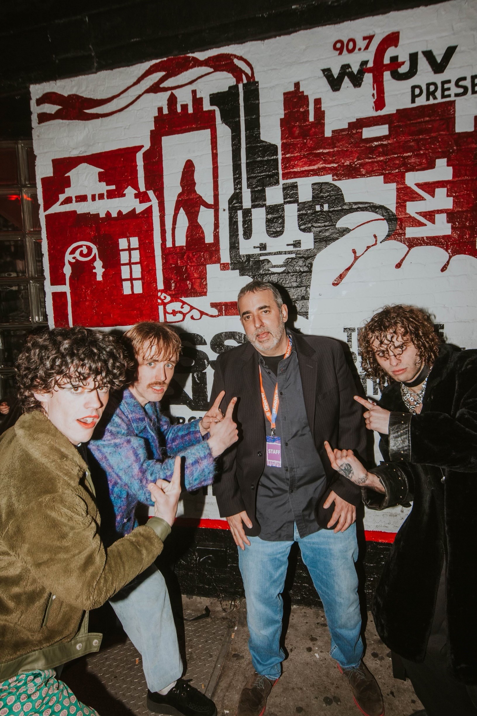 Four people standing in front of a red and black mural with buildings, a statue, and pipes, posing for the photo.