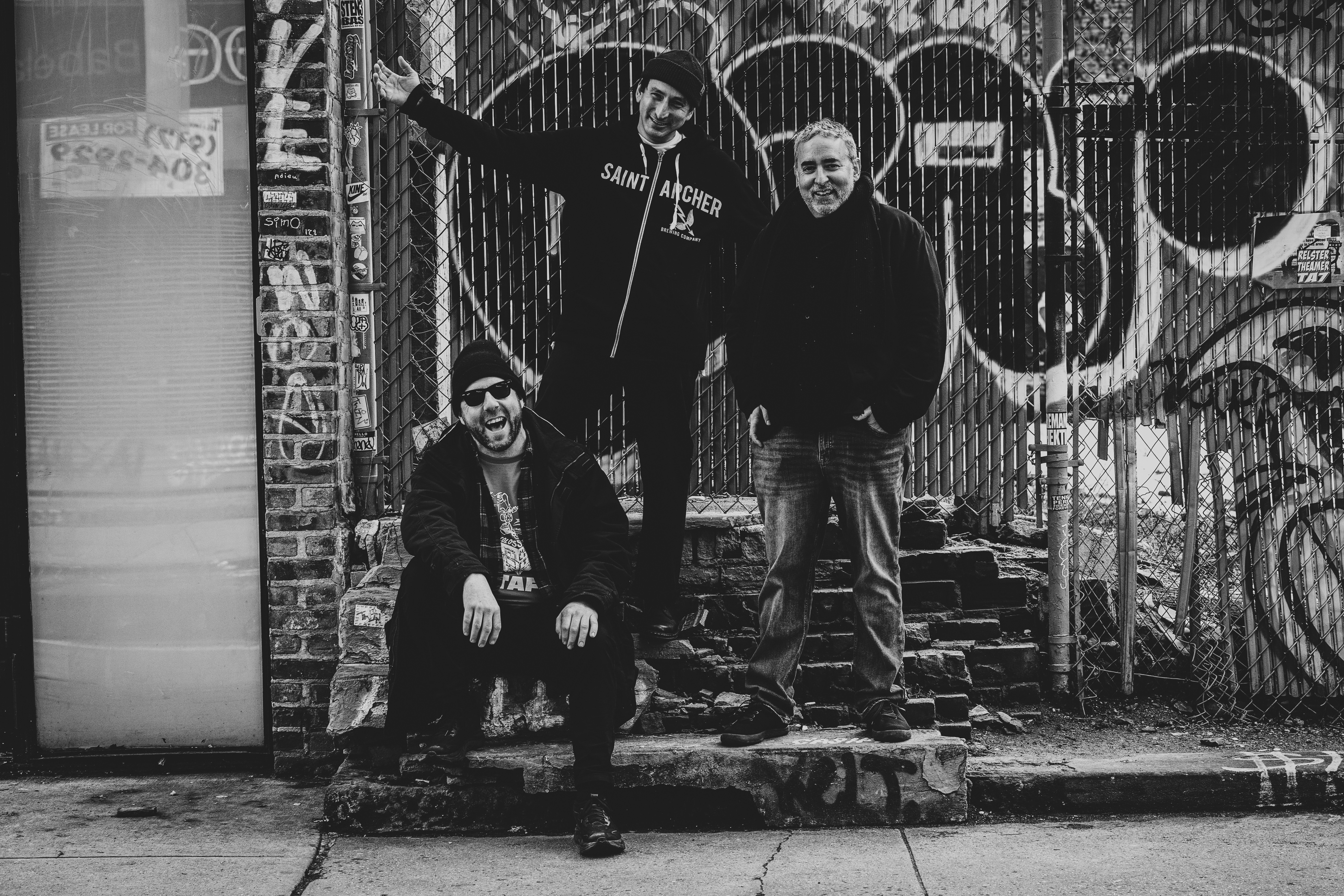 Three people pose in front of a graffiti-covered wall, with one person standing and two crouching, in black and white.