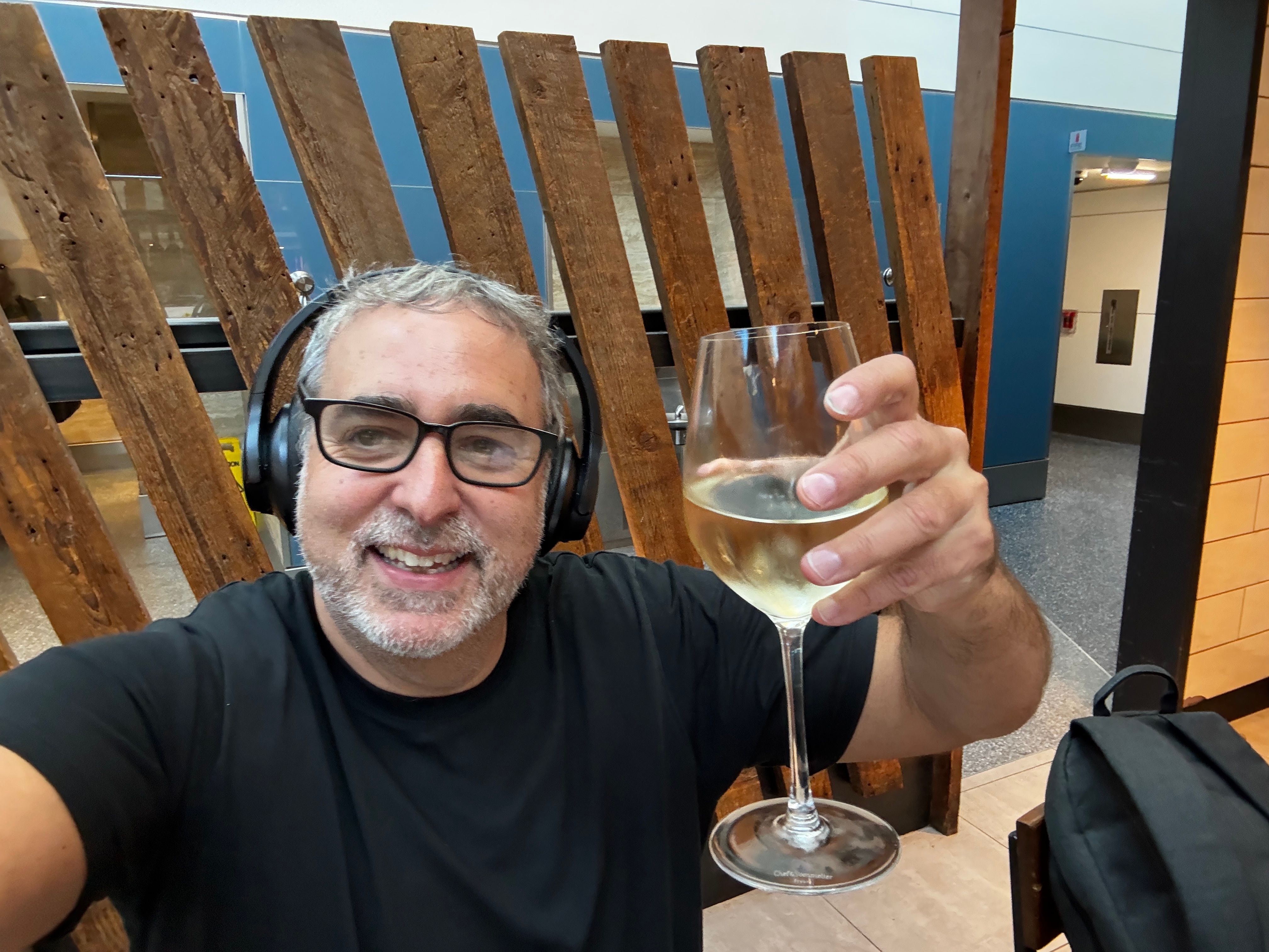 Man with glasses and headphones holding a glass of wine, smiling, sitting at a wooden table in a modern space.