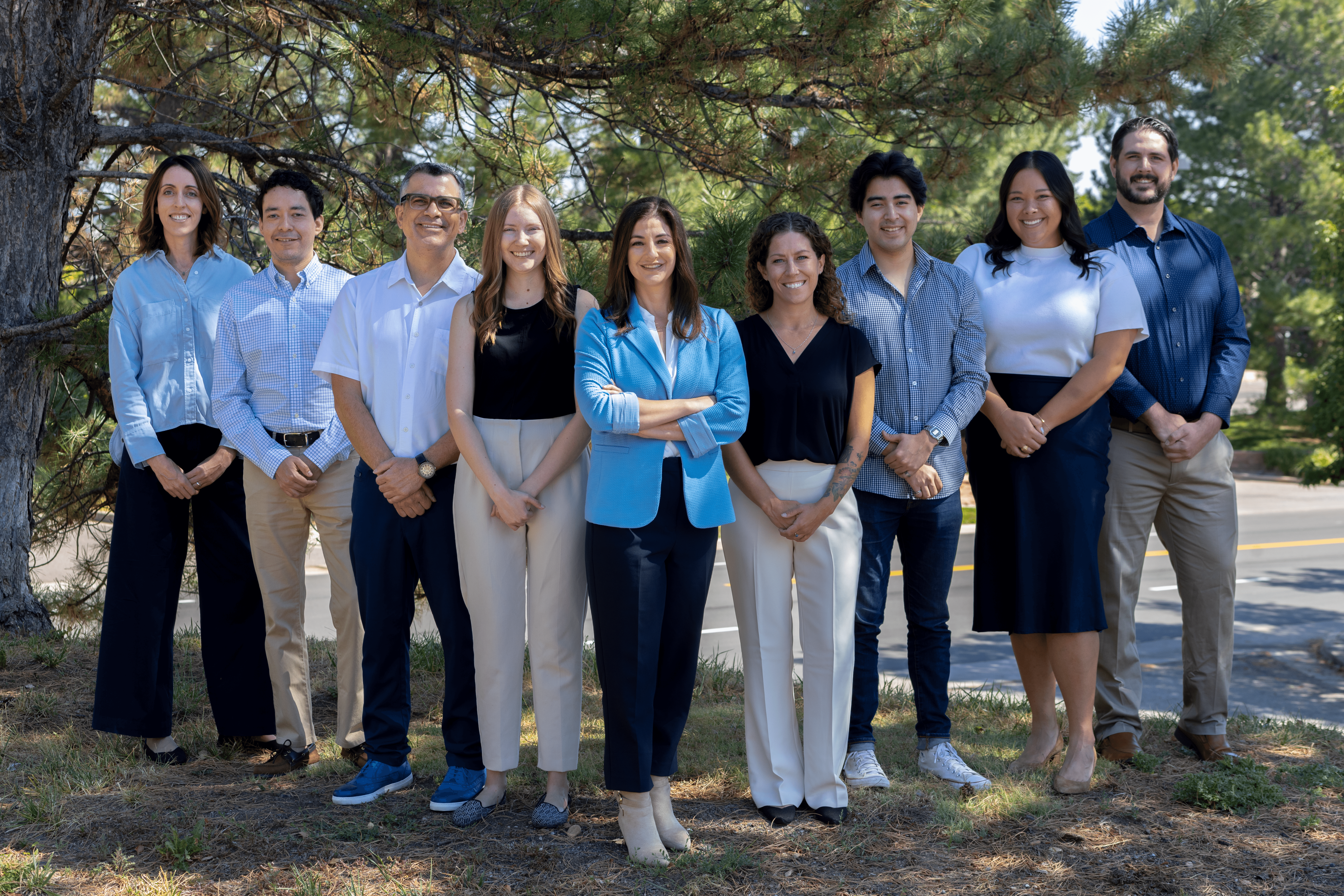 Group of nine people standing outdoors in front of trees, smiling, dressed in casual and business casual clothing.