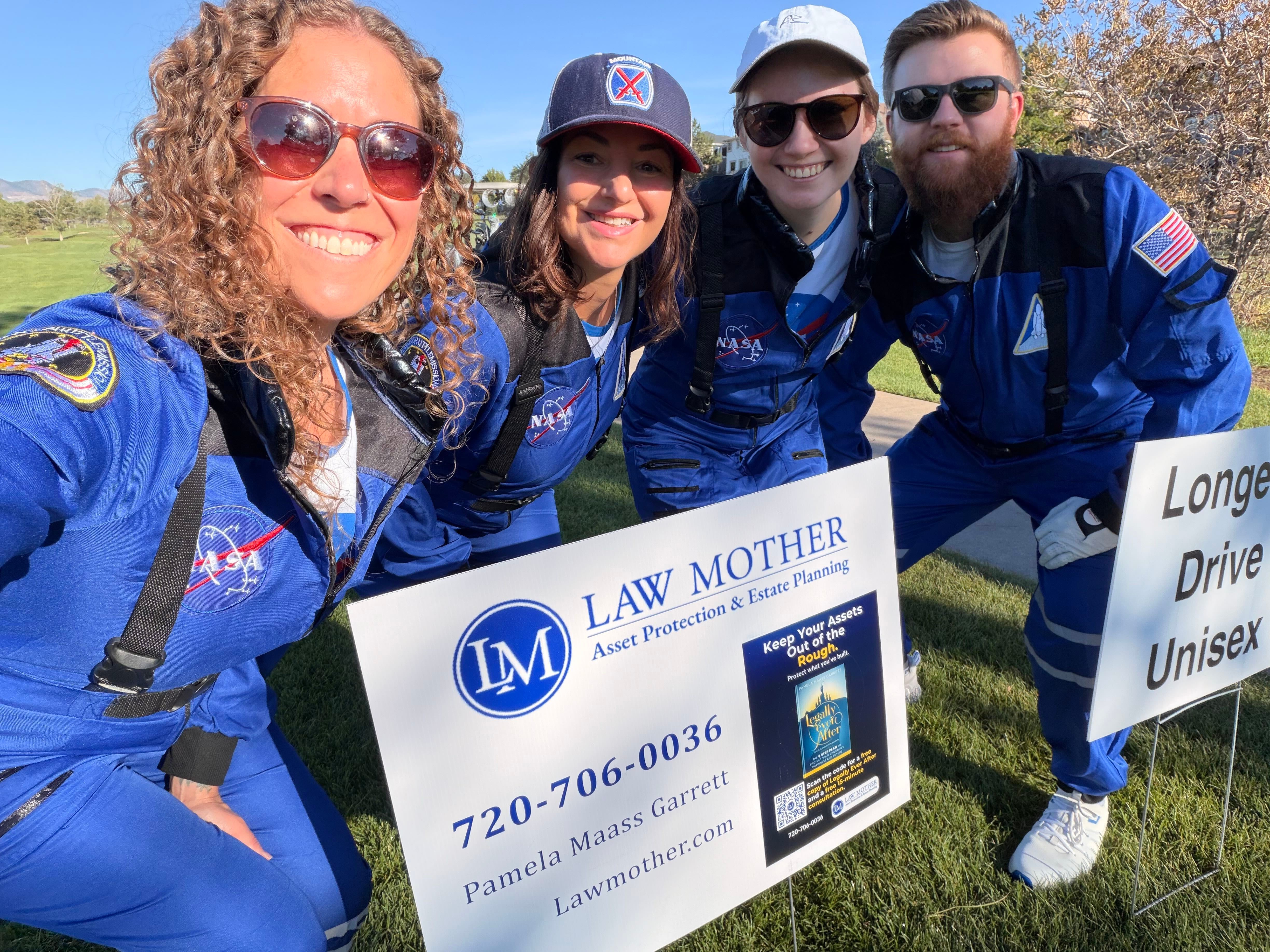 Four people in blue space suits smiling outdoors, holding a sign for law mother and a phone number.