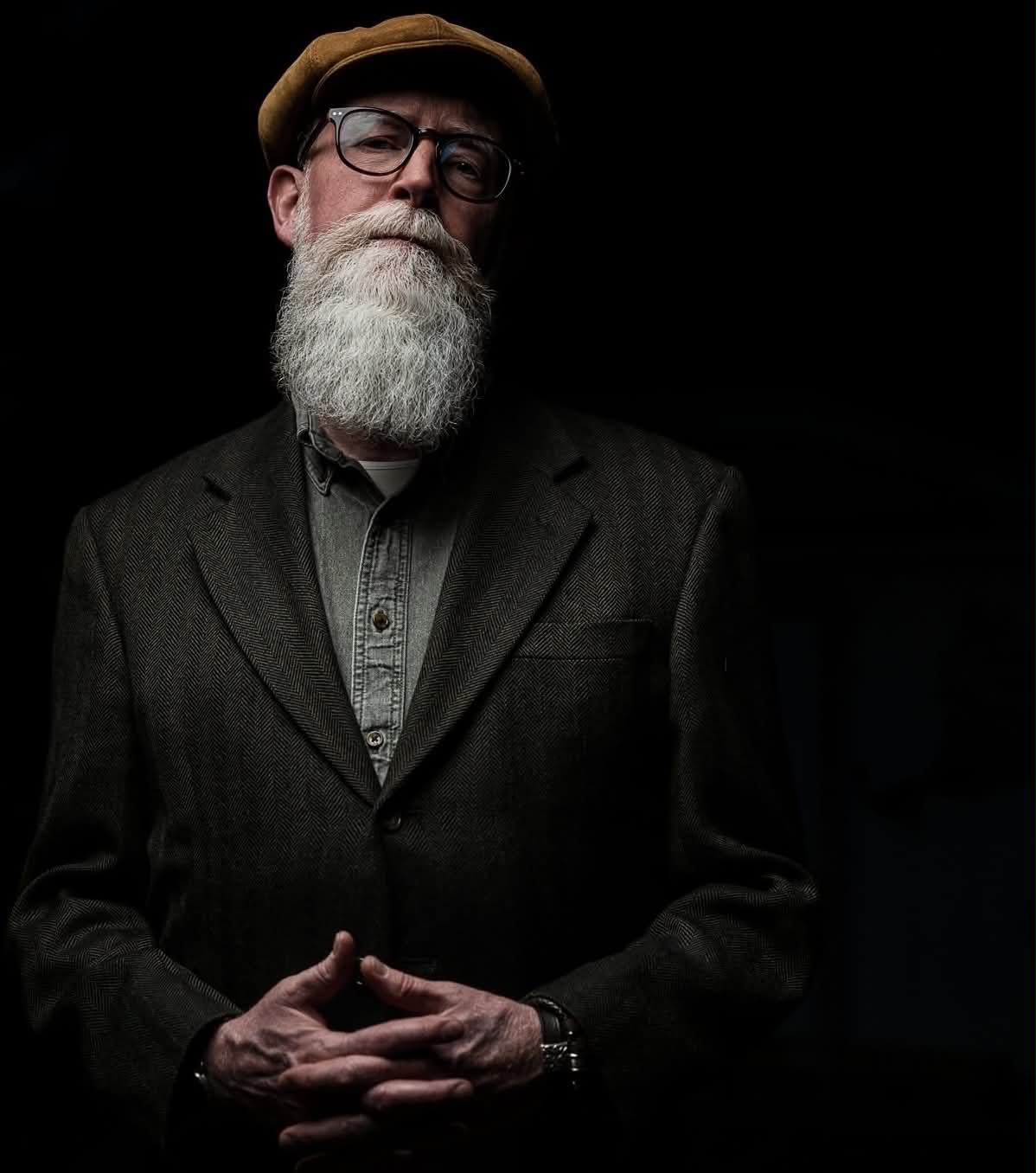 A bearded man wearing glasses, a hat, and a dark blazer, standing against a black background.