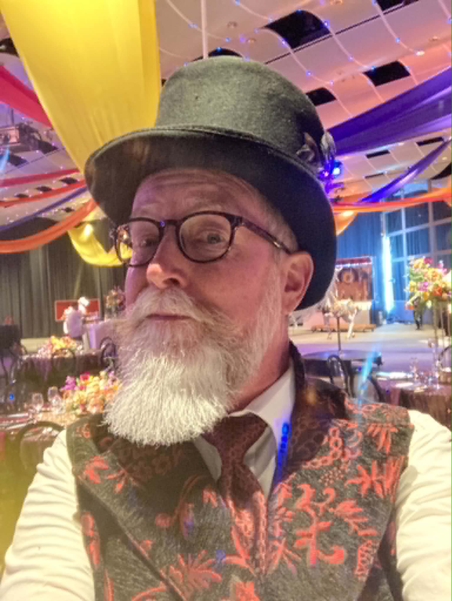 Man with glasses, gray beard, wearing a top hat and patterned vest, in a colorful decorated indoor setting.