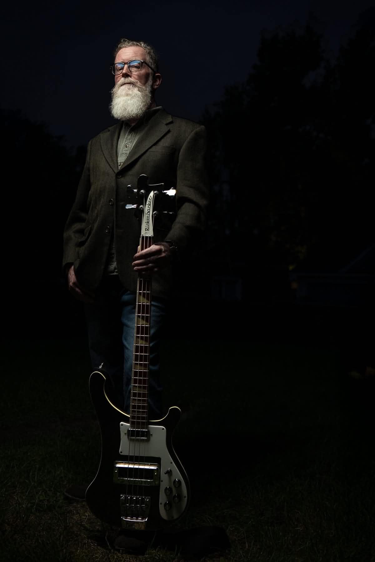 Man with a beard and glasses holding a guitar outdoors at night, dark background, standing with a serious expression.
