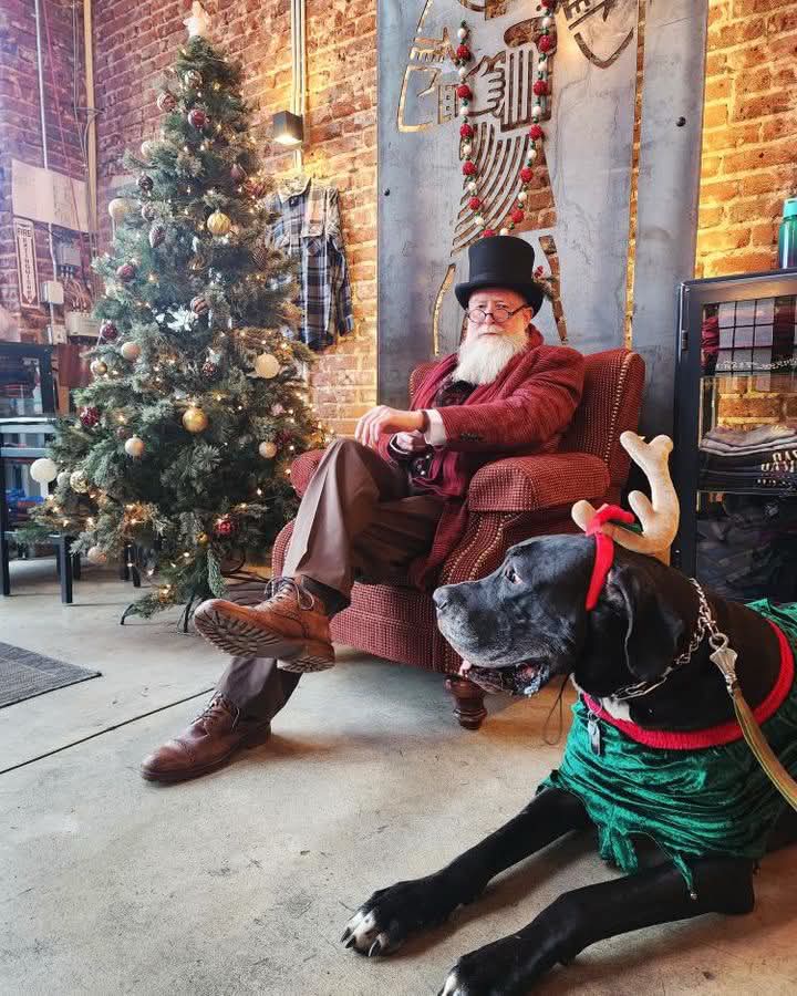 Man with a beard and glasses sitting in a chair, wearing a top hat and red sweater, with a black dog in festive clothing nearby, Christmas tree in background.