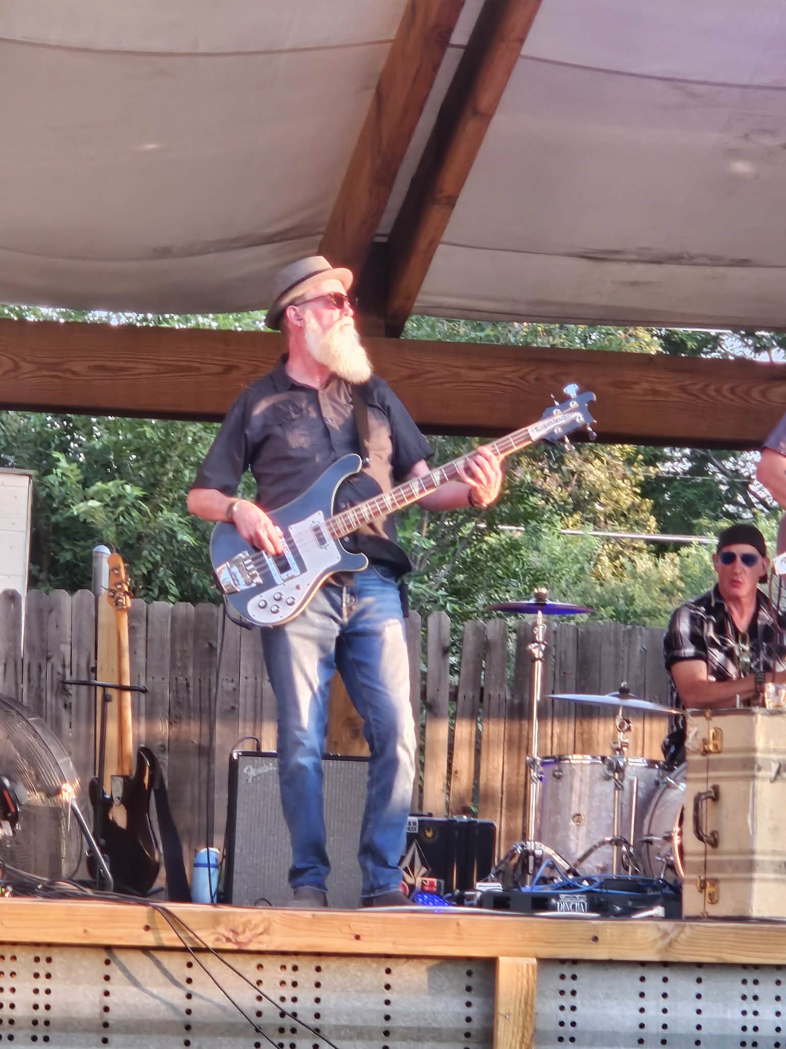 Man with a beard and sunglasses playing an electric guitar on stage outdoors, with another person in the background, under a canopy.
