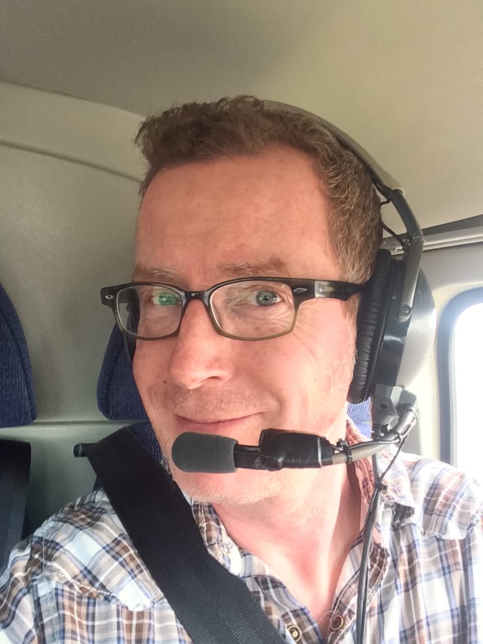 Man wearing glasses and a headset with microphone, smiling, inside vehicle, plaid shirt, seatbelt visible