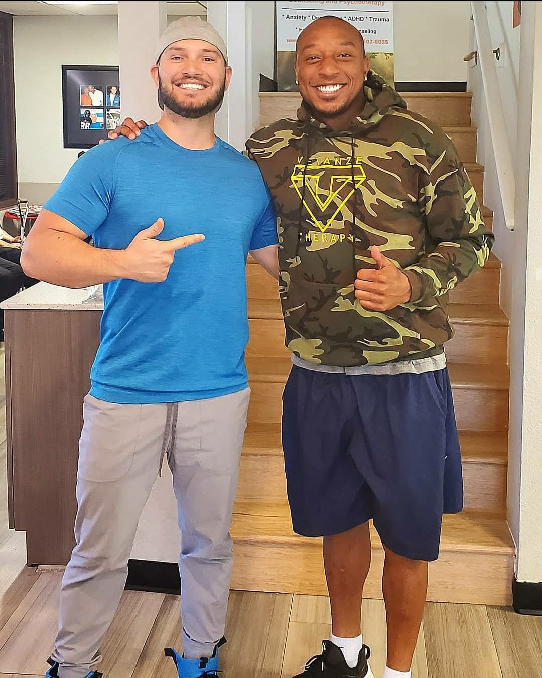 Two smiling men standing on stairs, one pointing at the other, in casual clothing indoors.