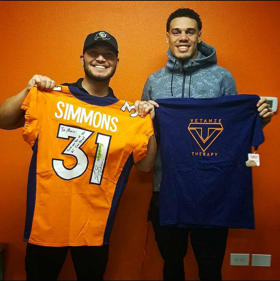 Two smiling men holding sports jerseys in front of an orange wall.