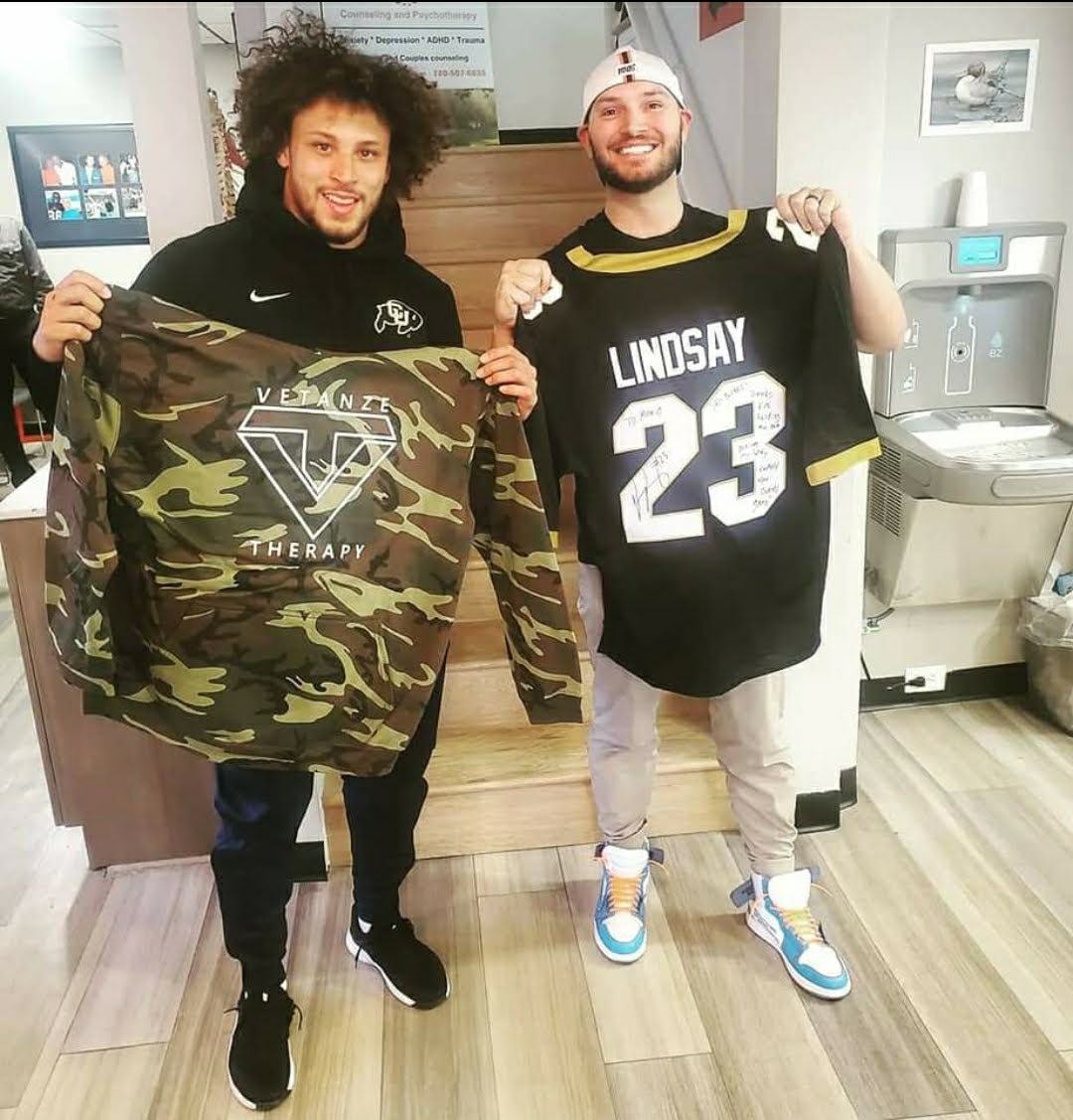 Two men smiling and holding sports jerseys indoors, one in camouflage and the other in a black jersey with the number 23.