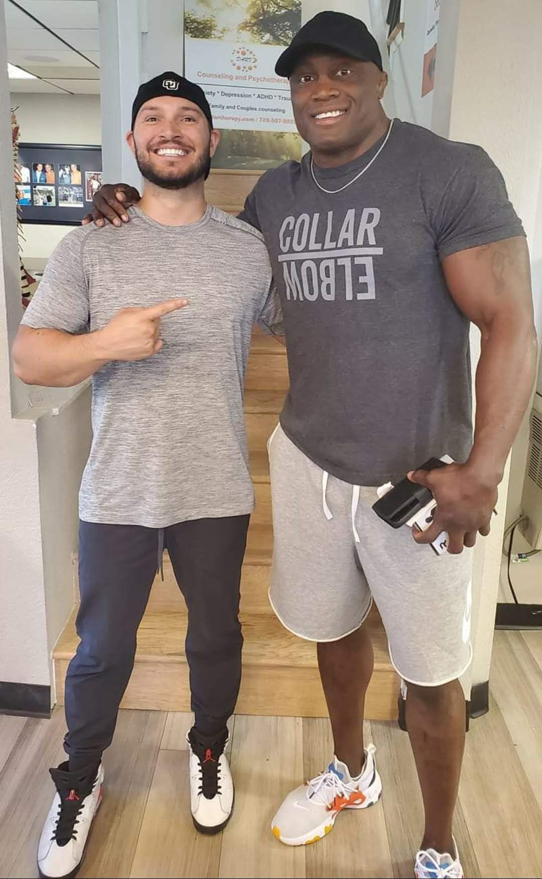 Two men standing together, smiling, one pointing at the other, in casual clothing indoors.