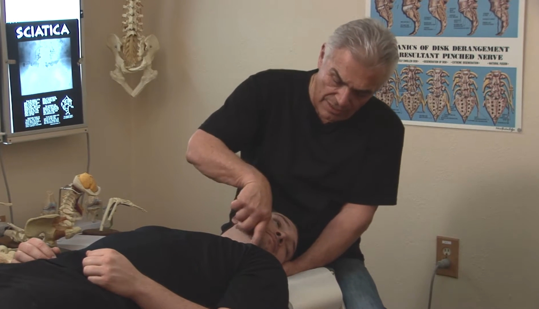 A person receives a chiropractic adjustment from a practitioner in a clinic room with anatomical posters and a skeleton model.