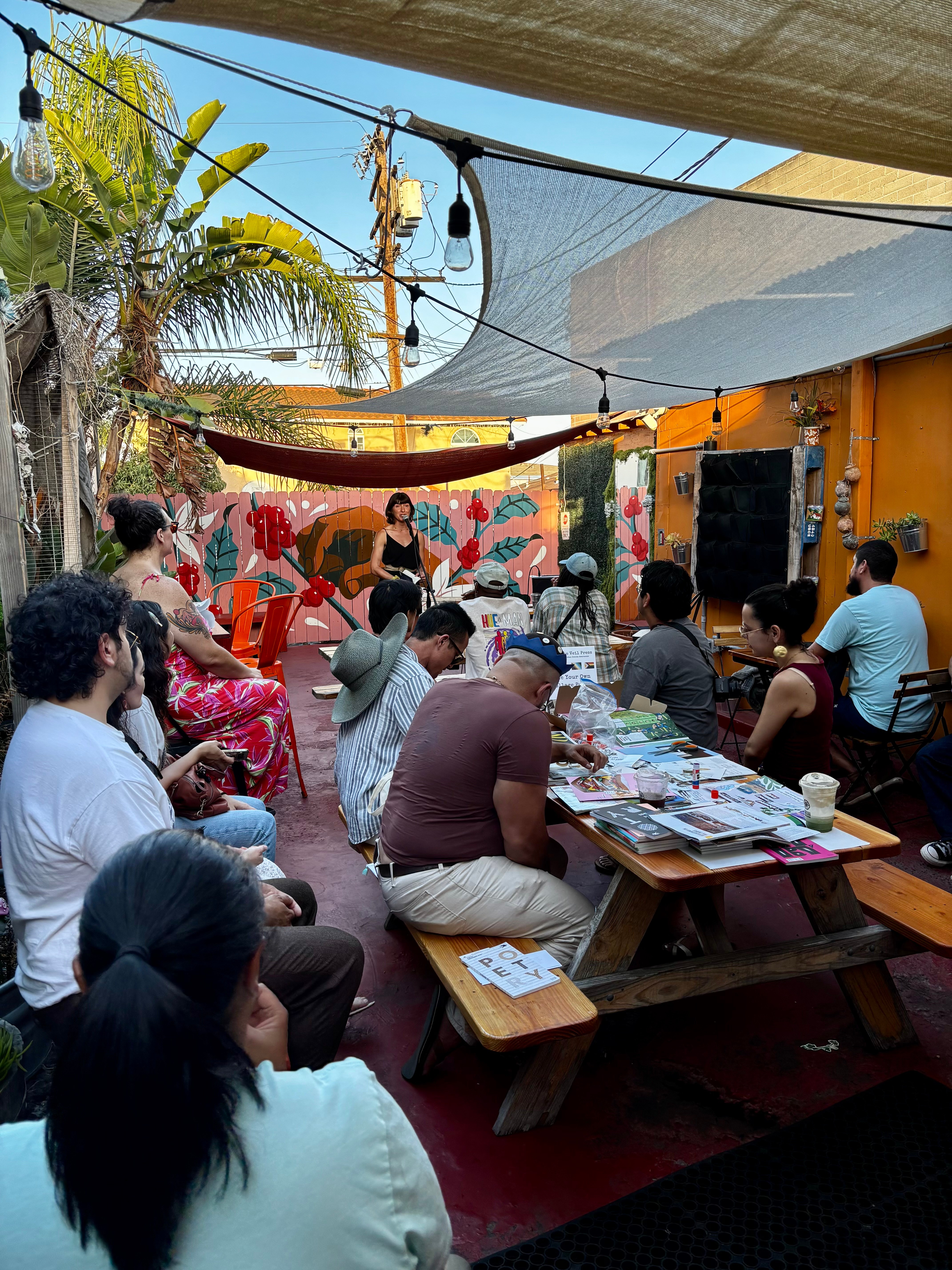 People gathered outdoors under a canopy, sitting at tables, with a performer on stage and colorful decorations in background.