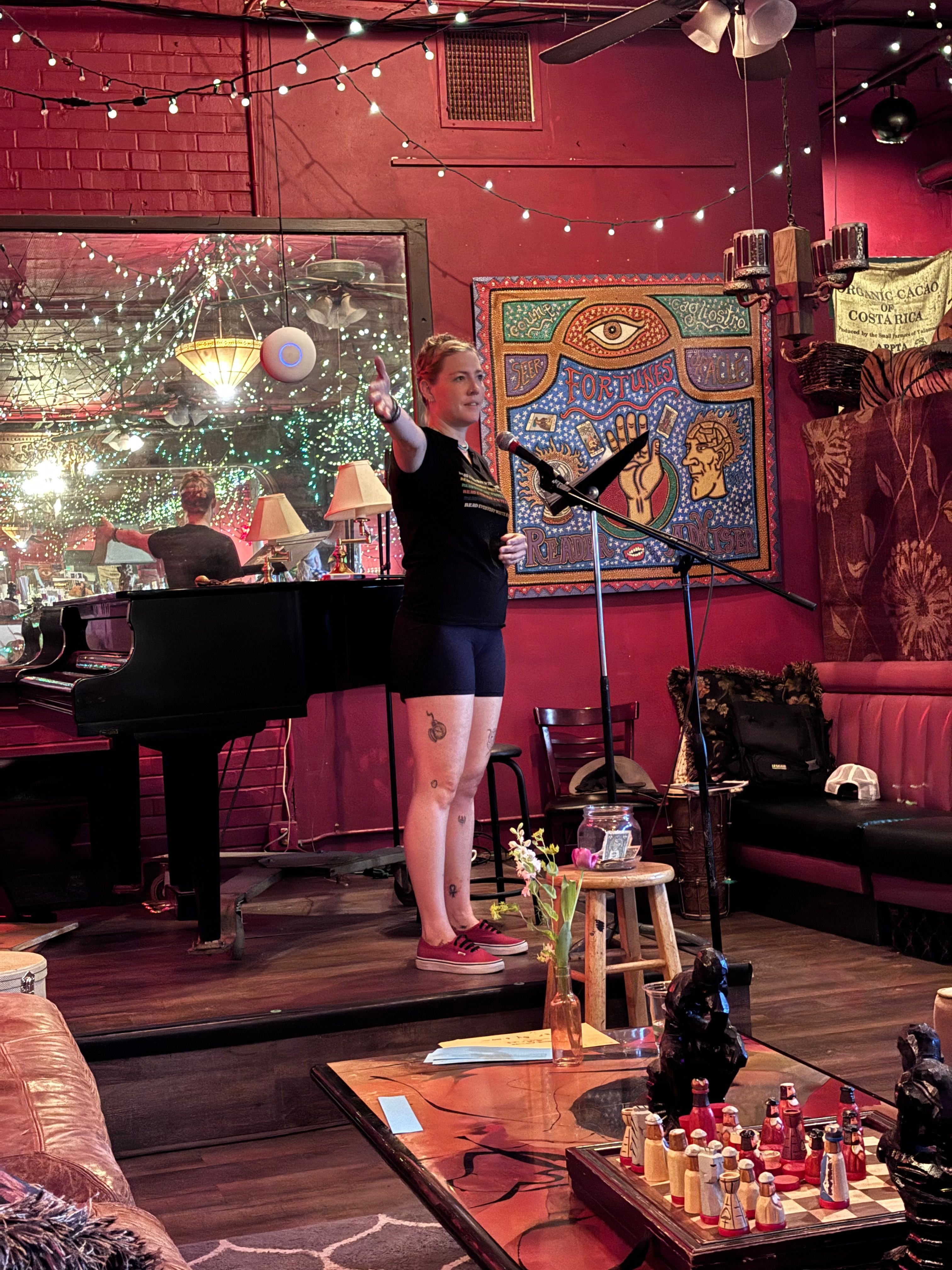 Two women on stage, one singing into a microphone, the other playing piano, in a decorated room with red walls and artwork.