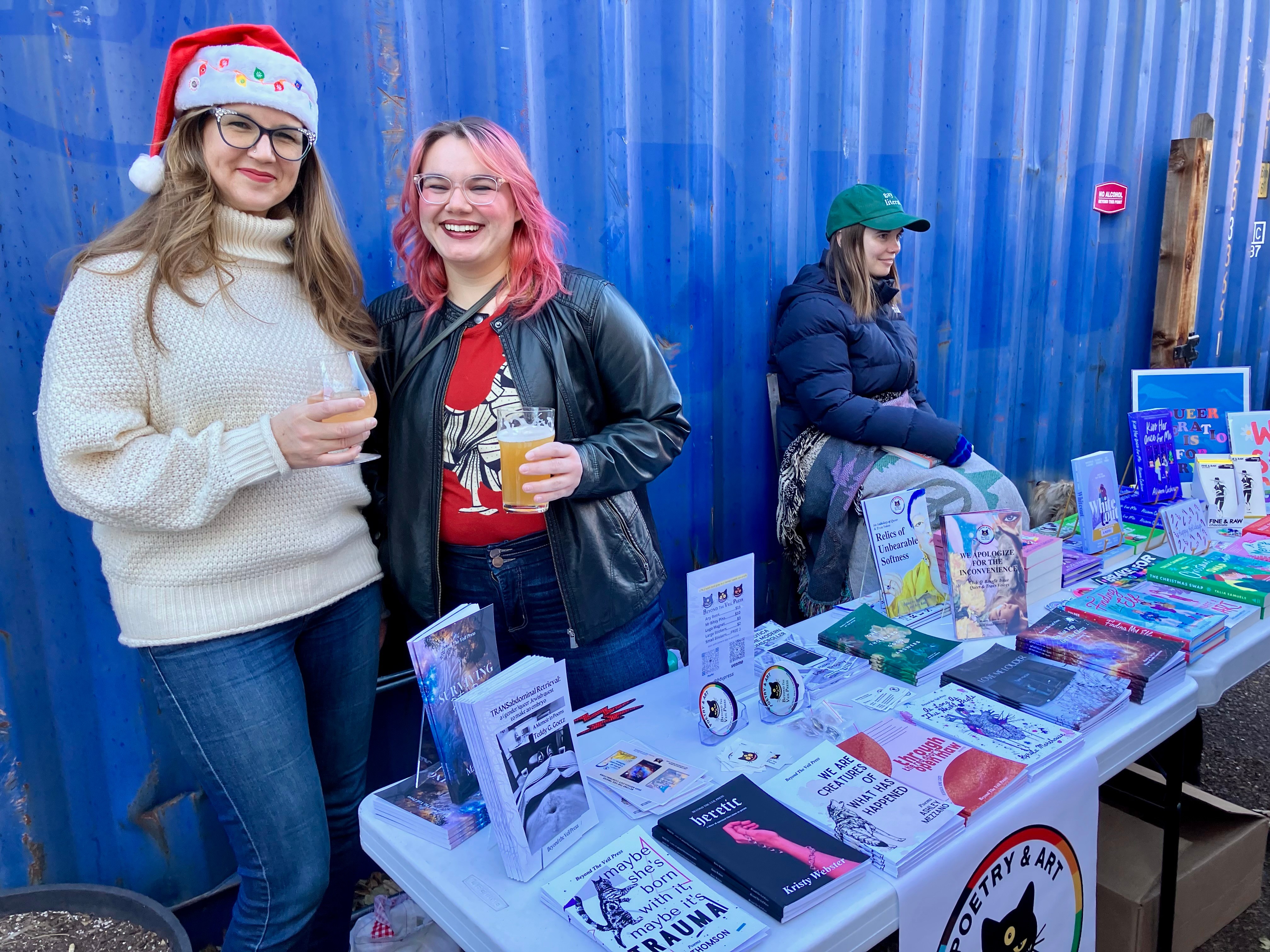 Two women smiling and holding drinks at a table with books and holiday decorations, one wearing a Santa hat, outdoors.