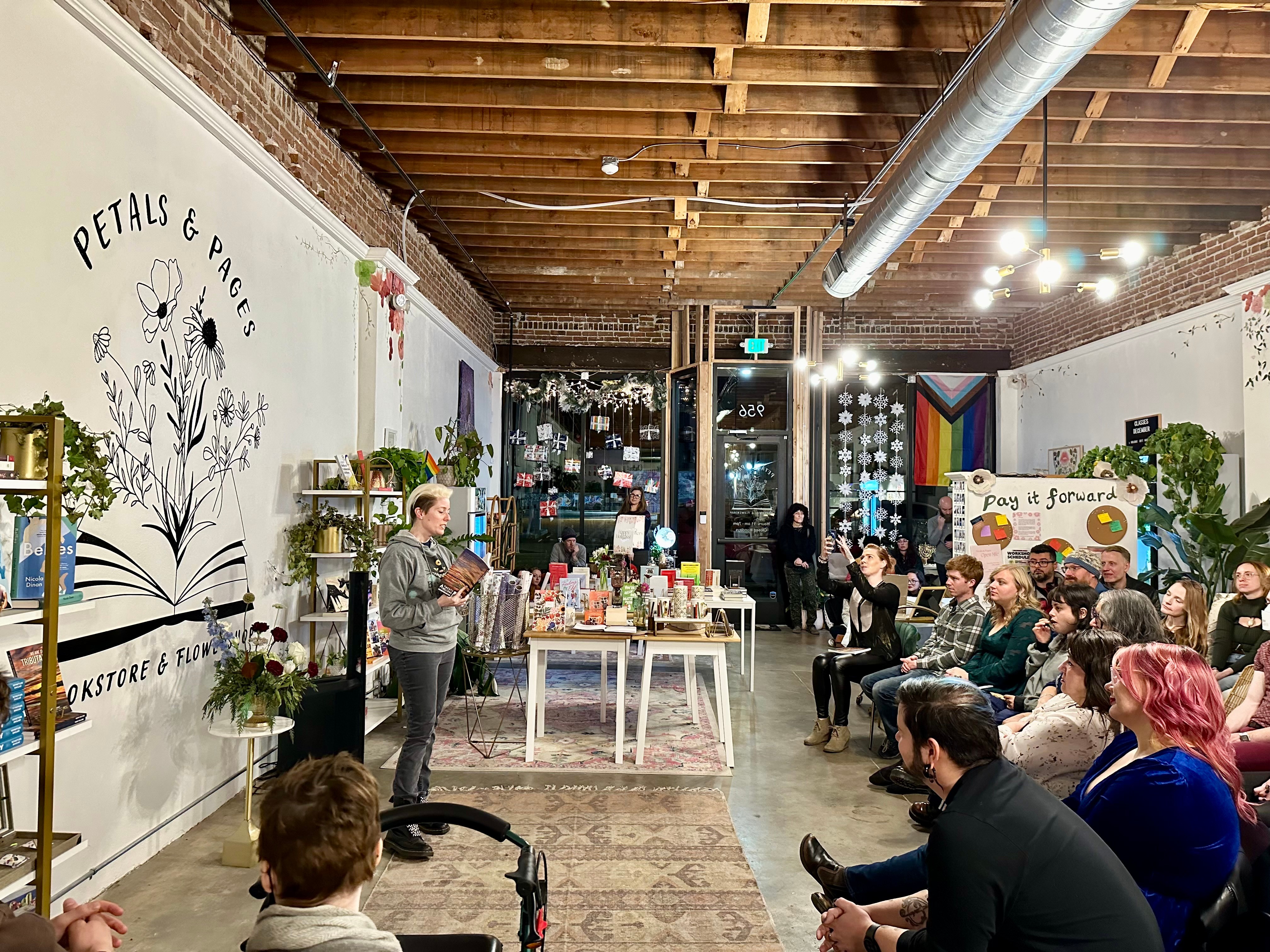 People gathered in a bookstore or event space with bookshelves, plants, and a large open book mural on the wall, attending a presentation or workshop.