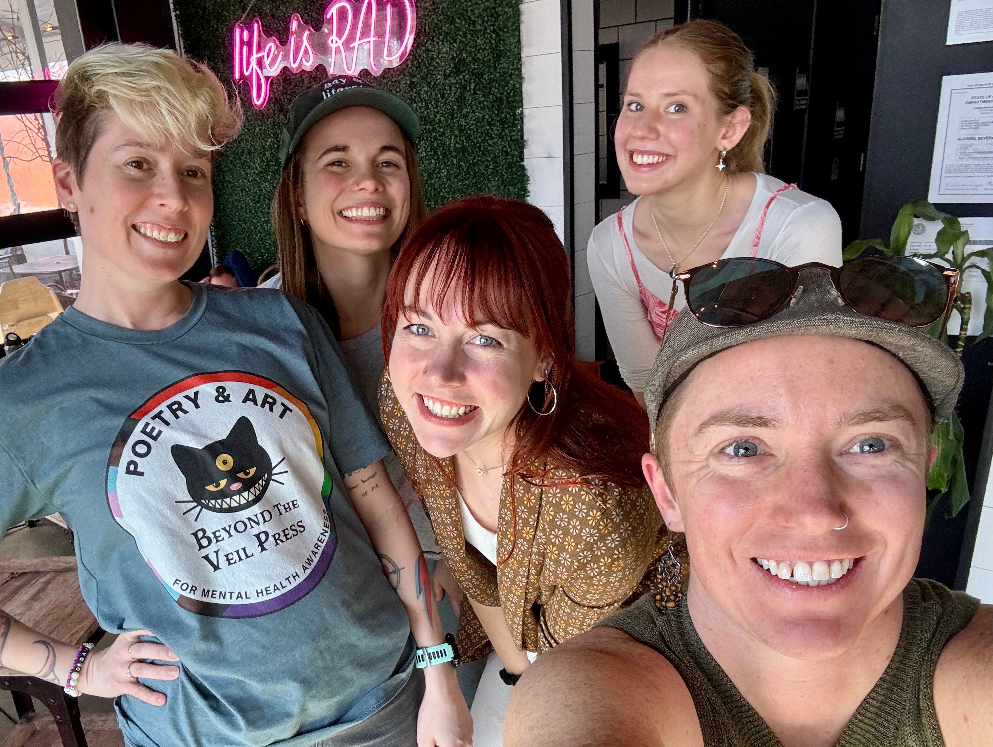 Five women smiling and posing for a selfie indoors with a neon sign in the background.