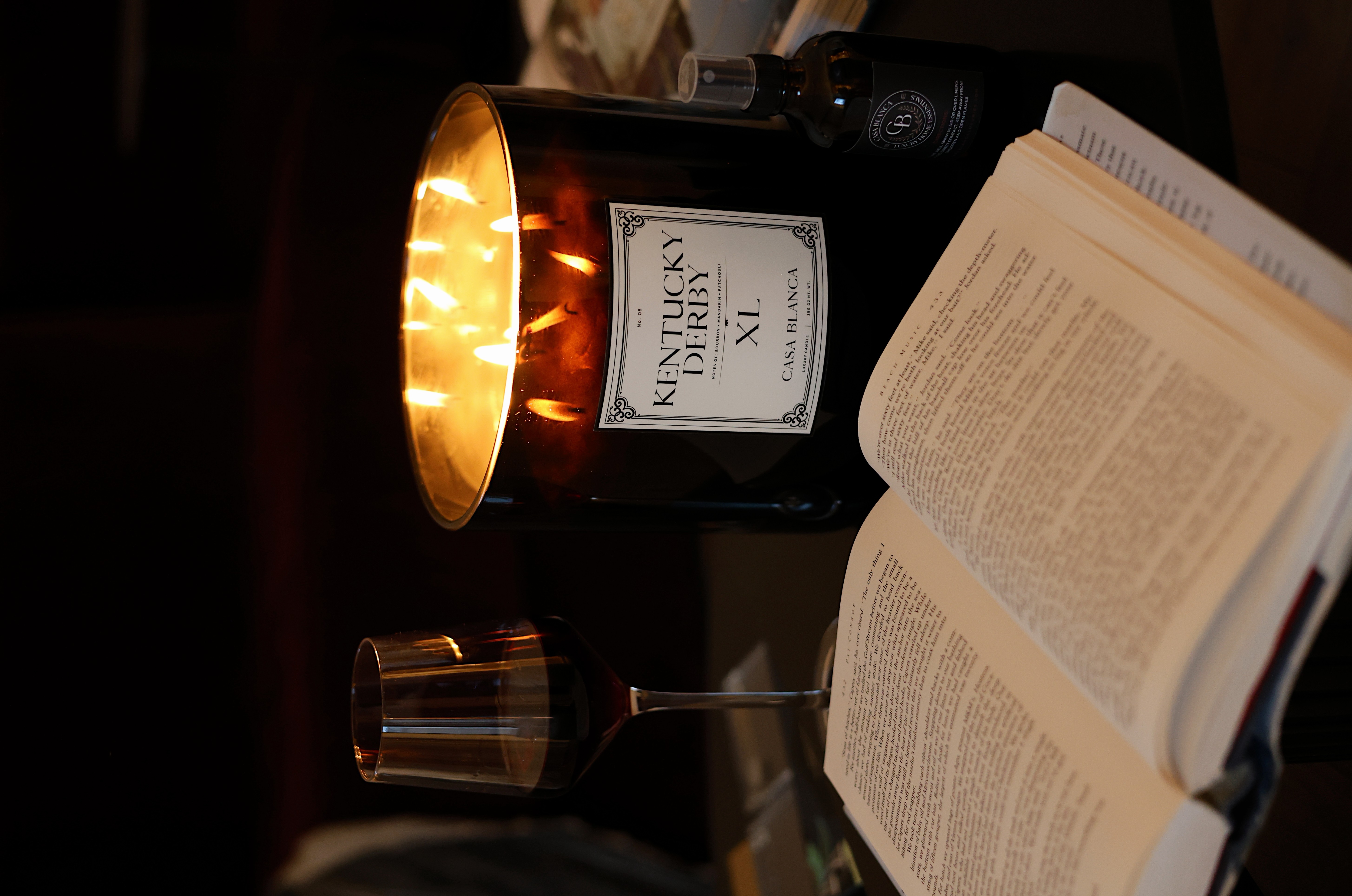 Lit candle in a dark room, open book, and glass of wine on a table.