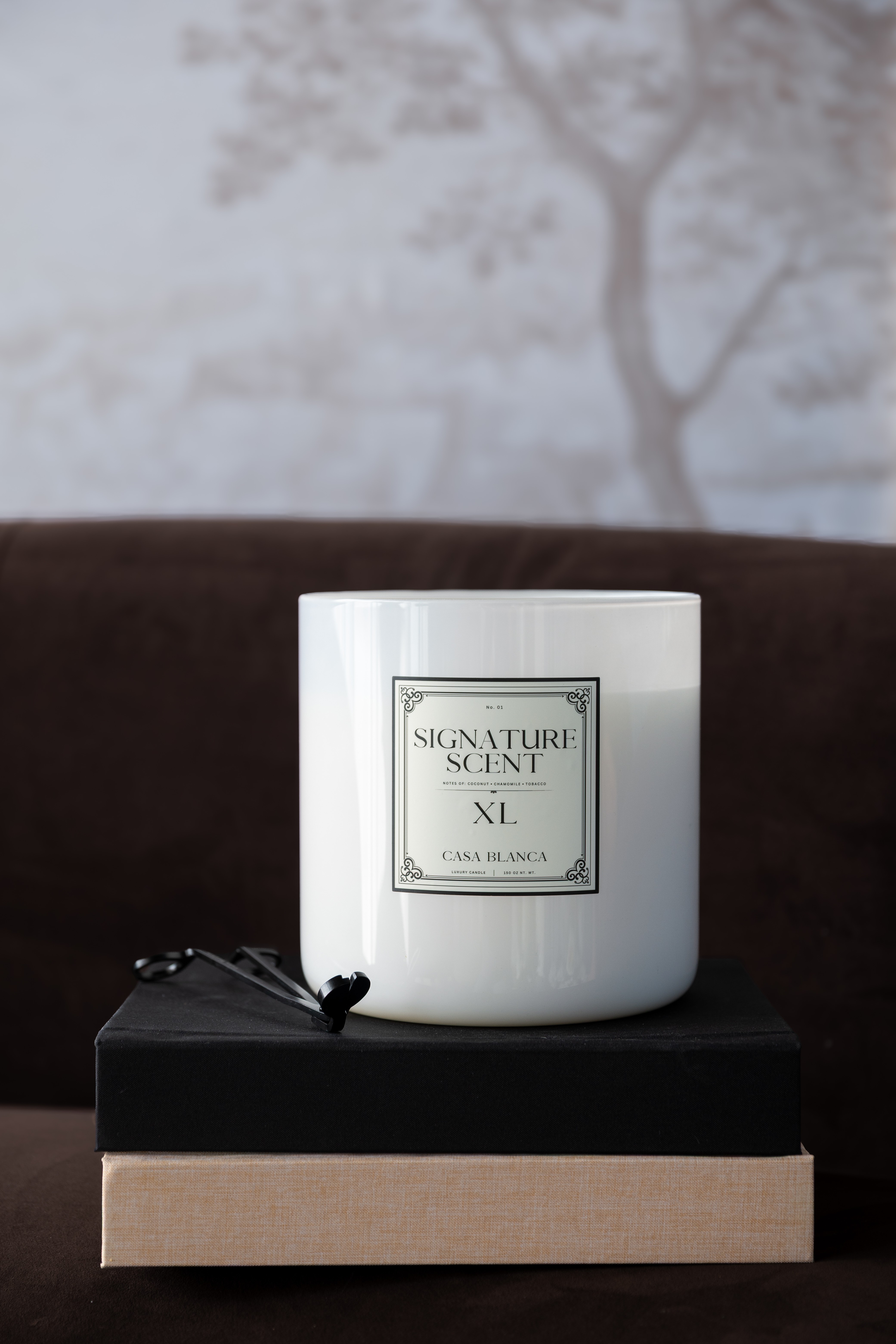 White scented candle on a black and beige box, with a blurred tree background, in a cozy setting.