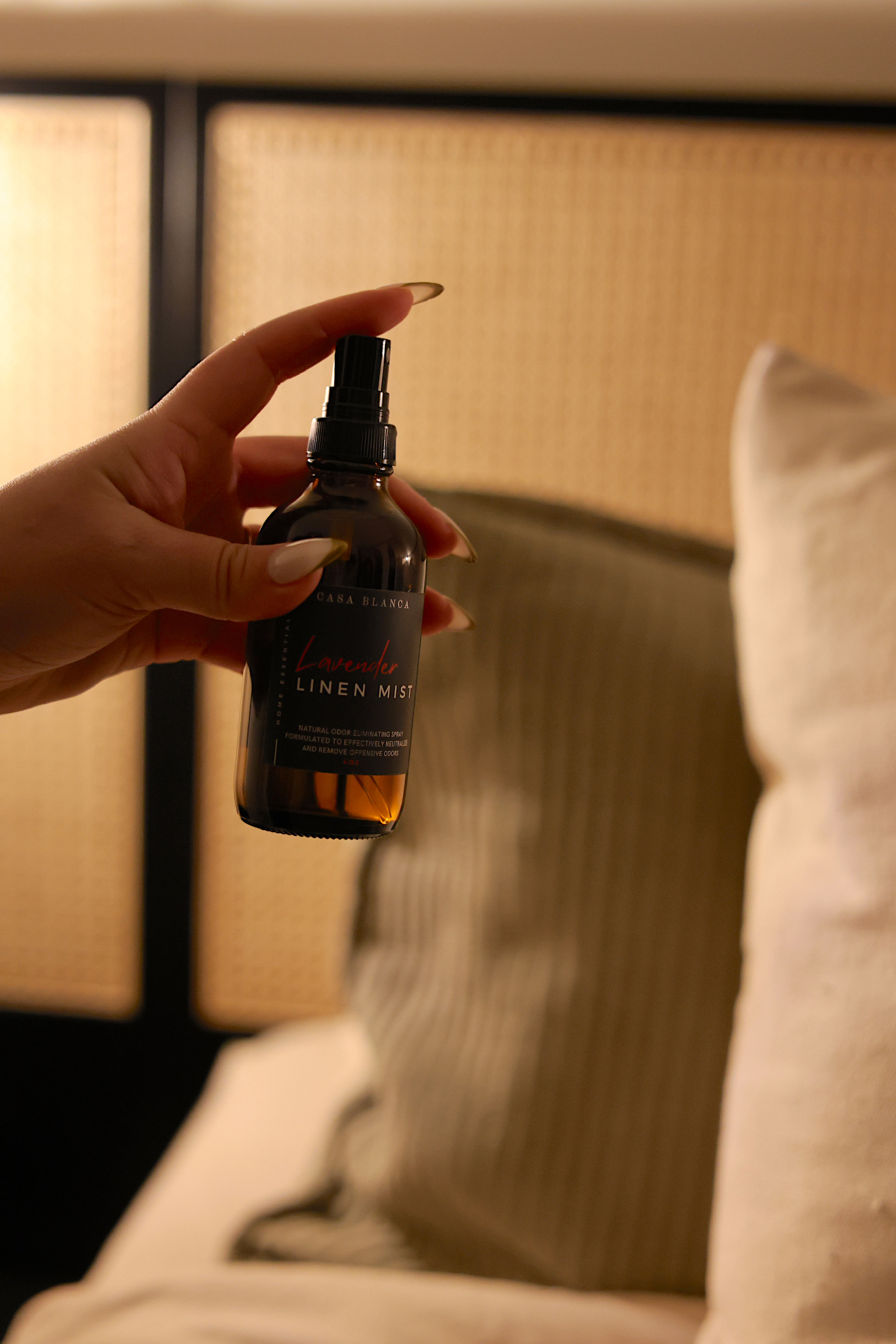 Hand holding a small spray bottle with a black cap, in front of a bed with beige pillows and headboard.