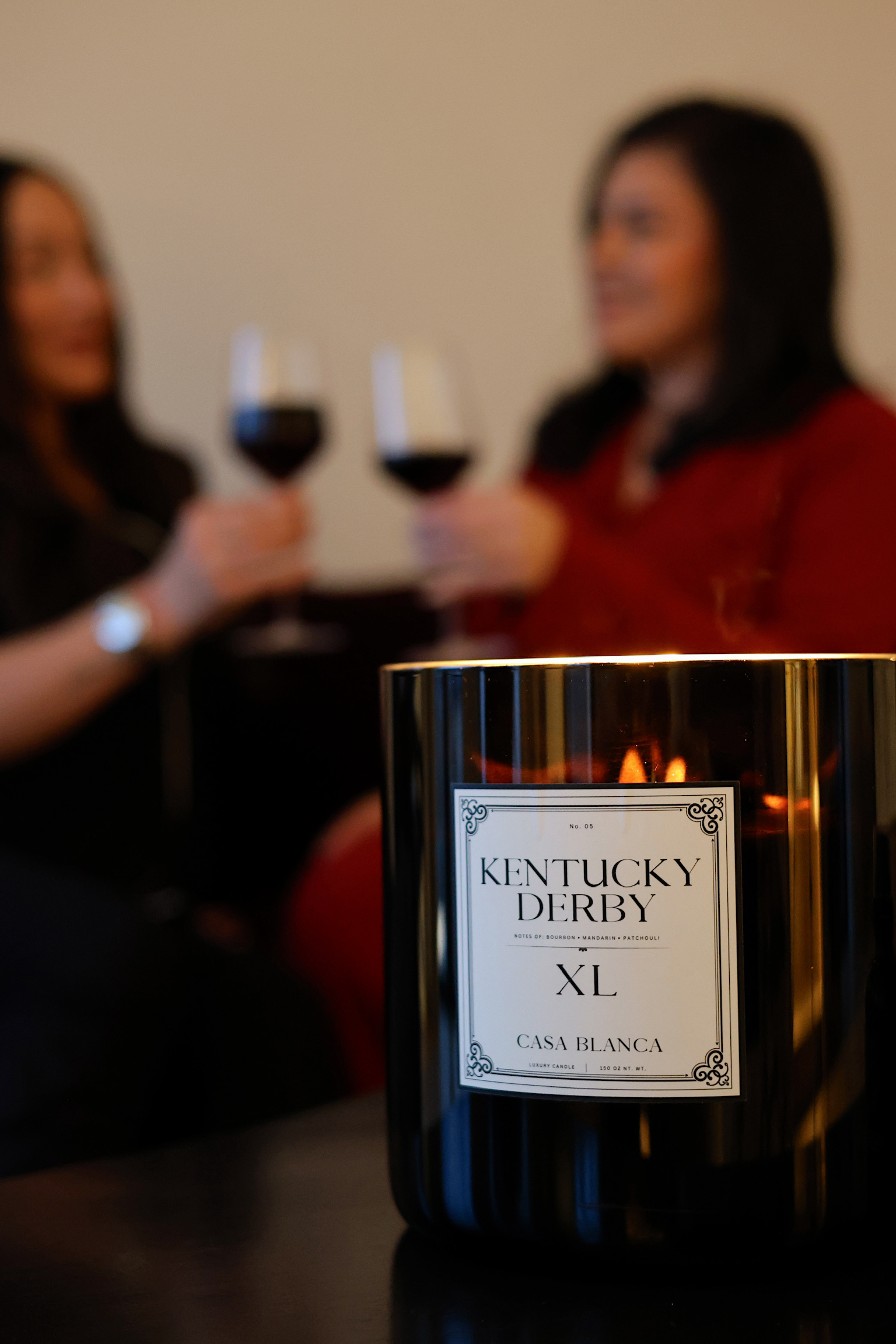 Two women holding glasses of red wine, with a candle and a black container labeled 'Kentucky Derby XL' in the foreground.