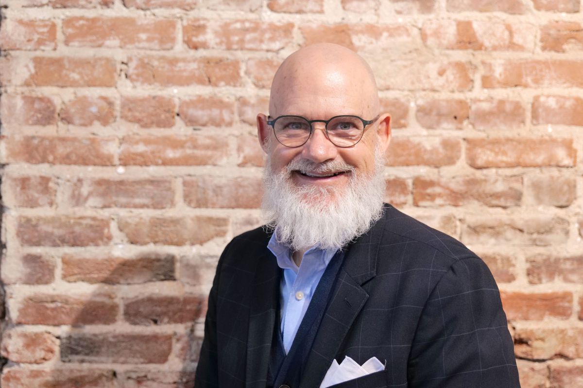 Bald man with glasses and a white beard smiling, wearing a dark blazer and light blue shirt, standing in front of a brick wall.