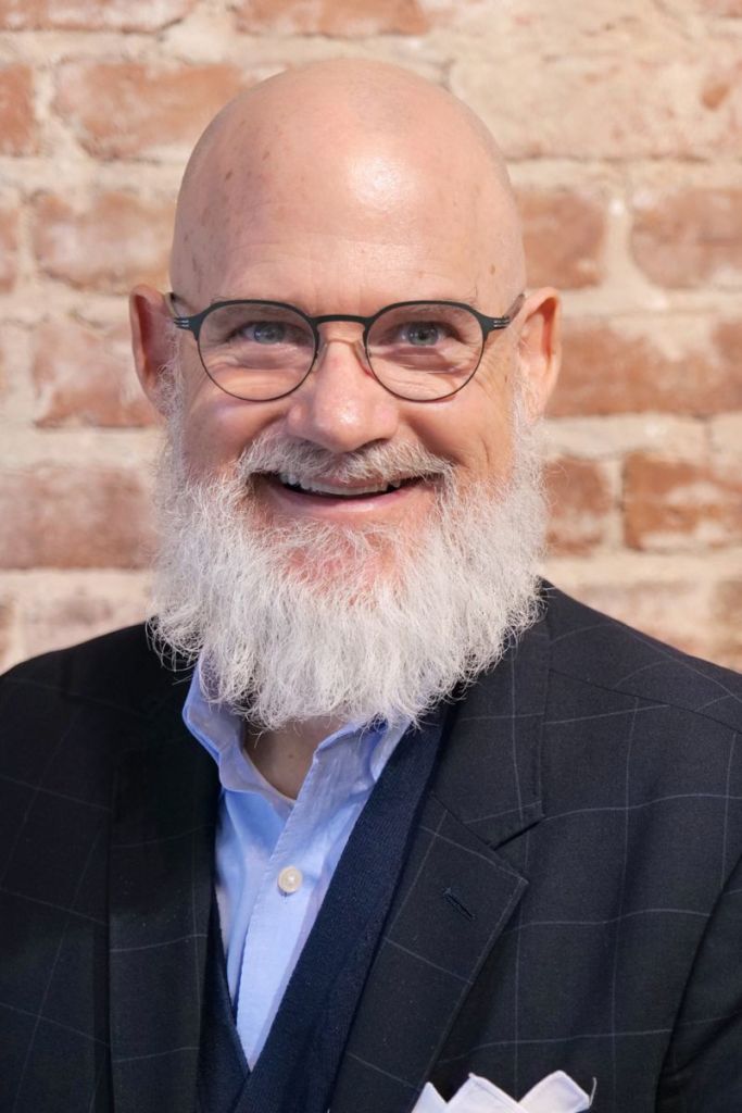 Bald man with glasses and a white beard smiling, wearing a dark blazer and light blue shirt, brick wall background.