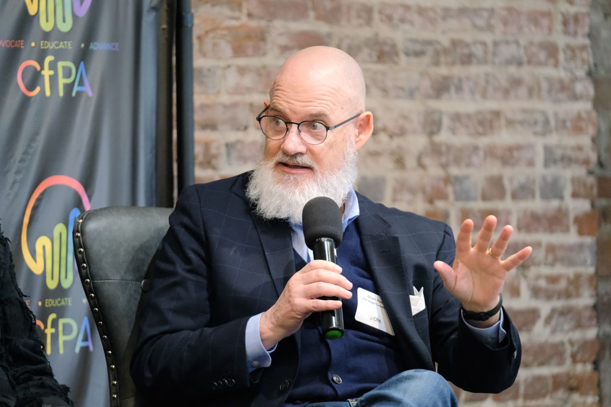 Bald man with glasses and a beard speaking into a microphone, gesturing with his left hand, seated indoors with a brick wall background.