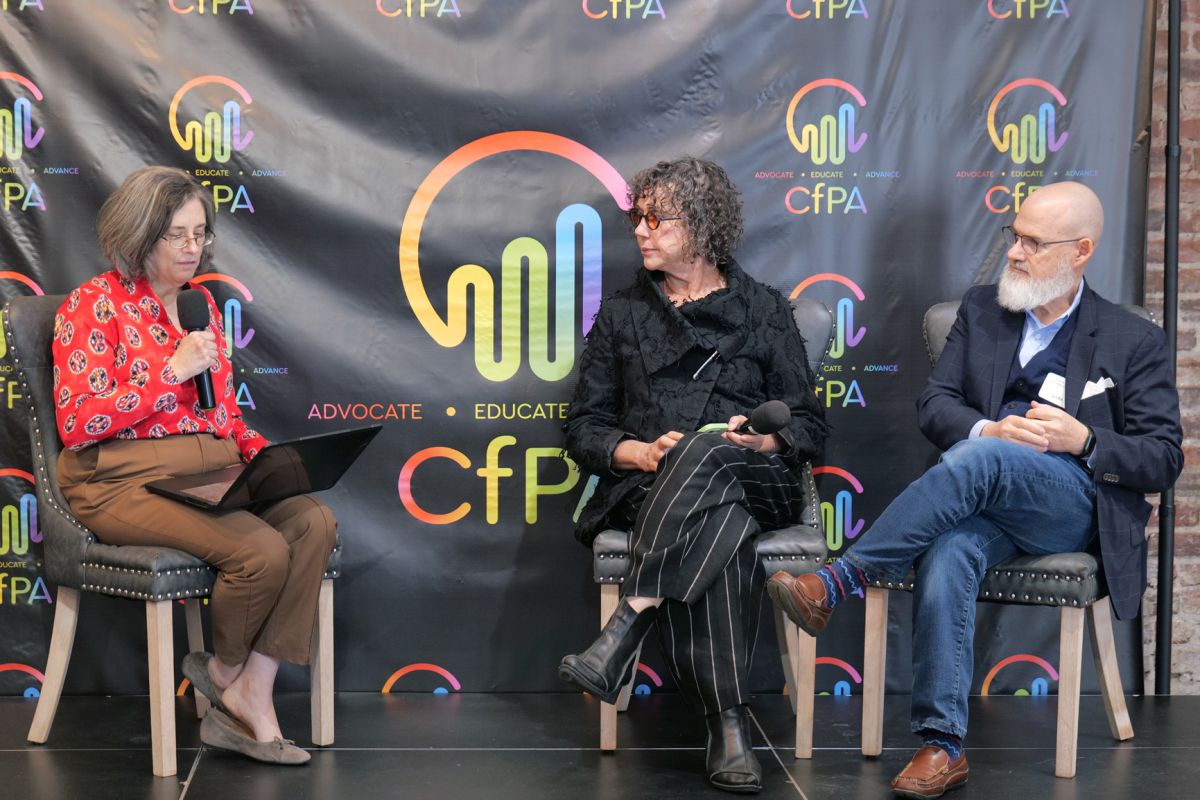 Three people seated on stage in front of a black backdrop with colorful logos, engaged in conversation during a panel discussion.