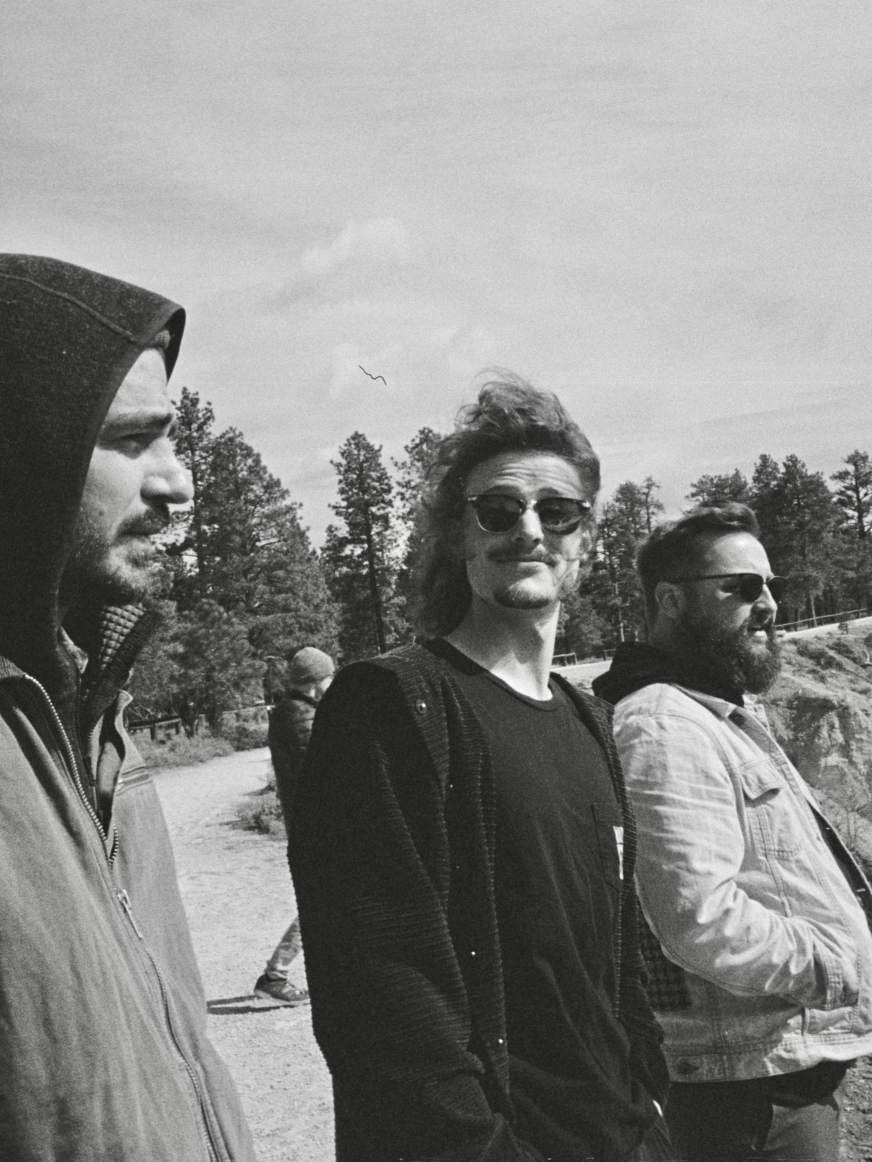 Three people standing outdoors with trees and sky in the background, one wearing sunglasses, in black and white.
