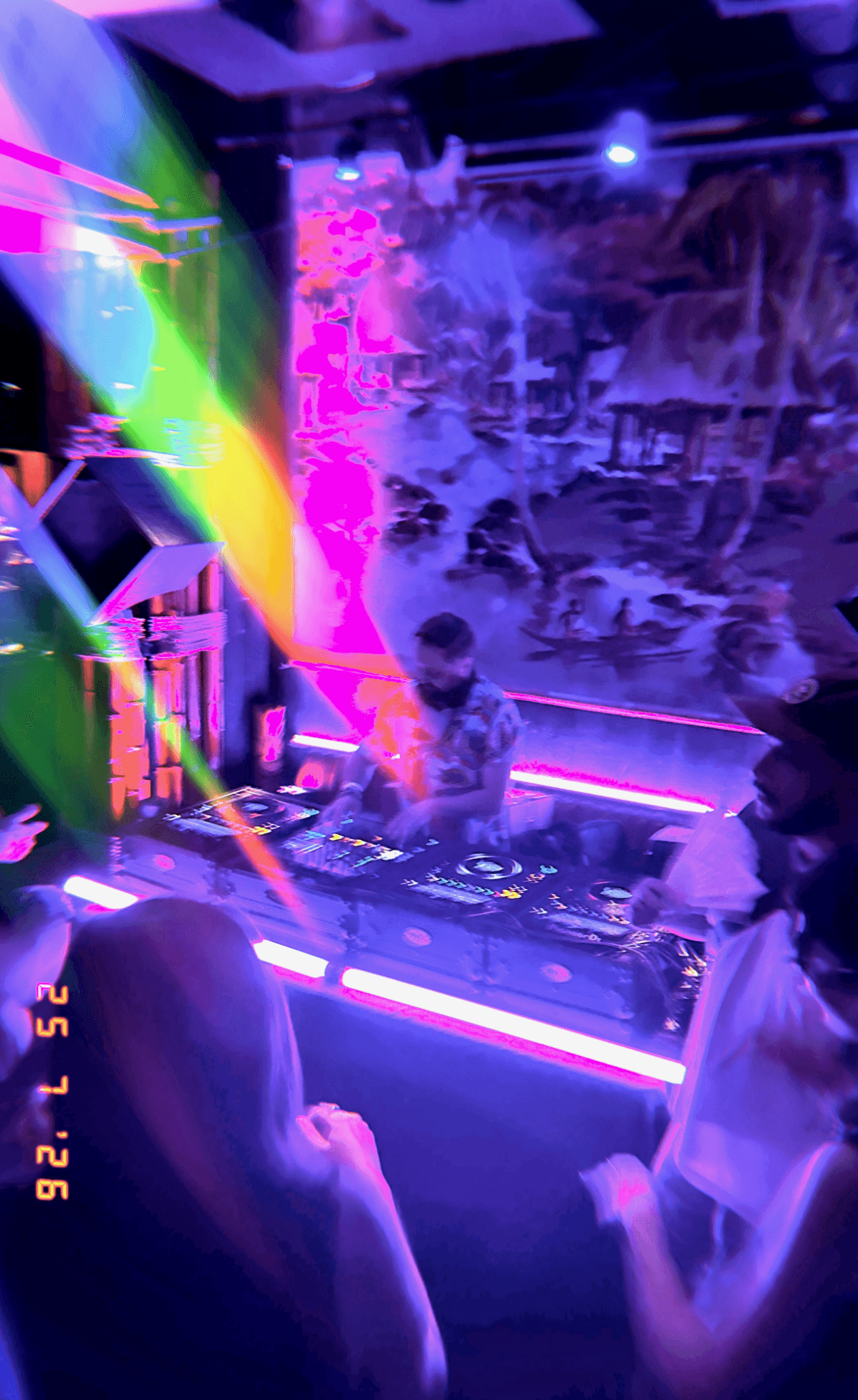 DJ performing at a colorful nightclub with vibrant lights and a foggy atmosphere, people dancing and enjoying the music.
