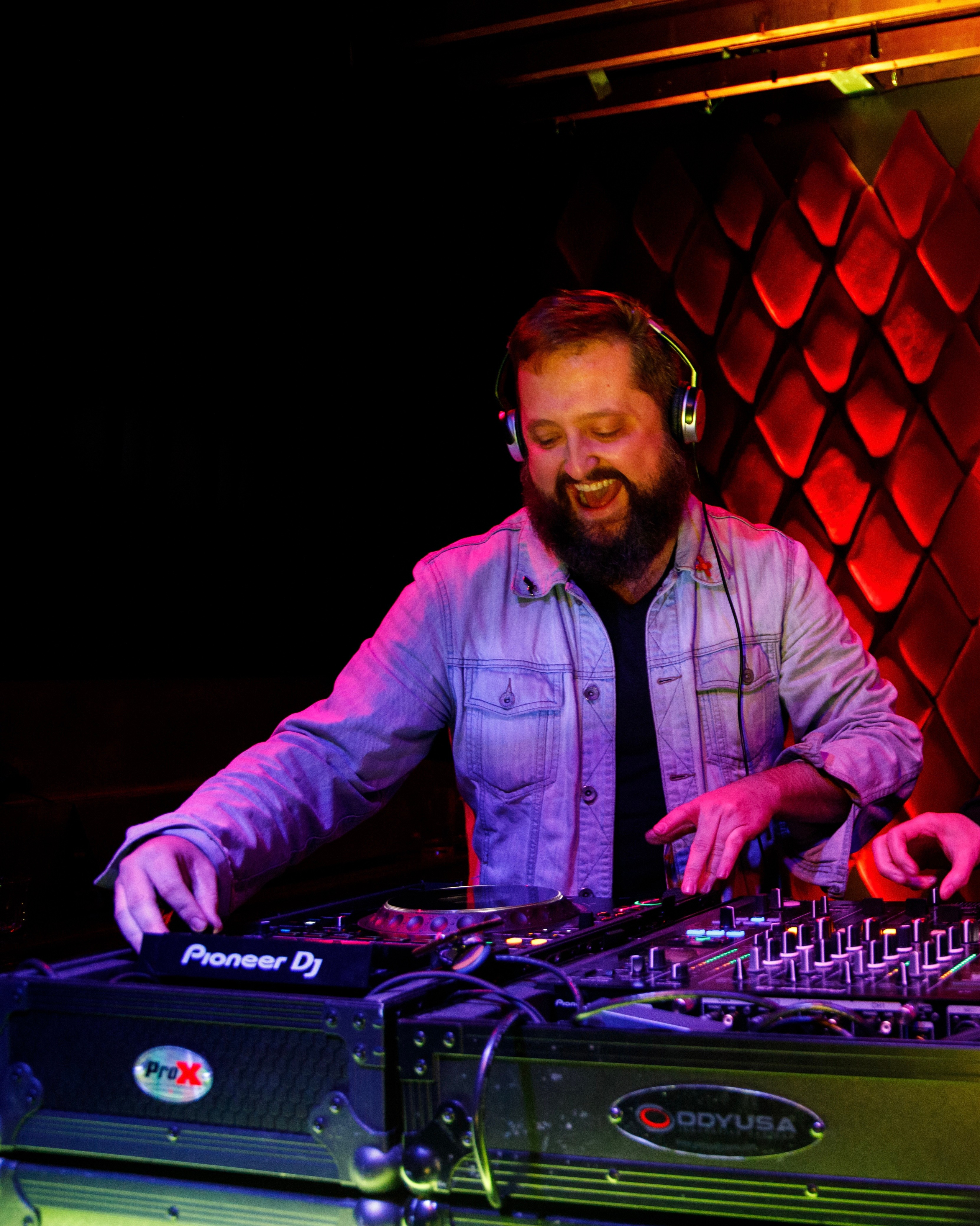 DJ with headphones operating a mixer and turntables in a dimly lit venue with red wall panels.